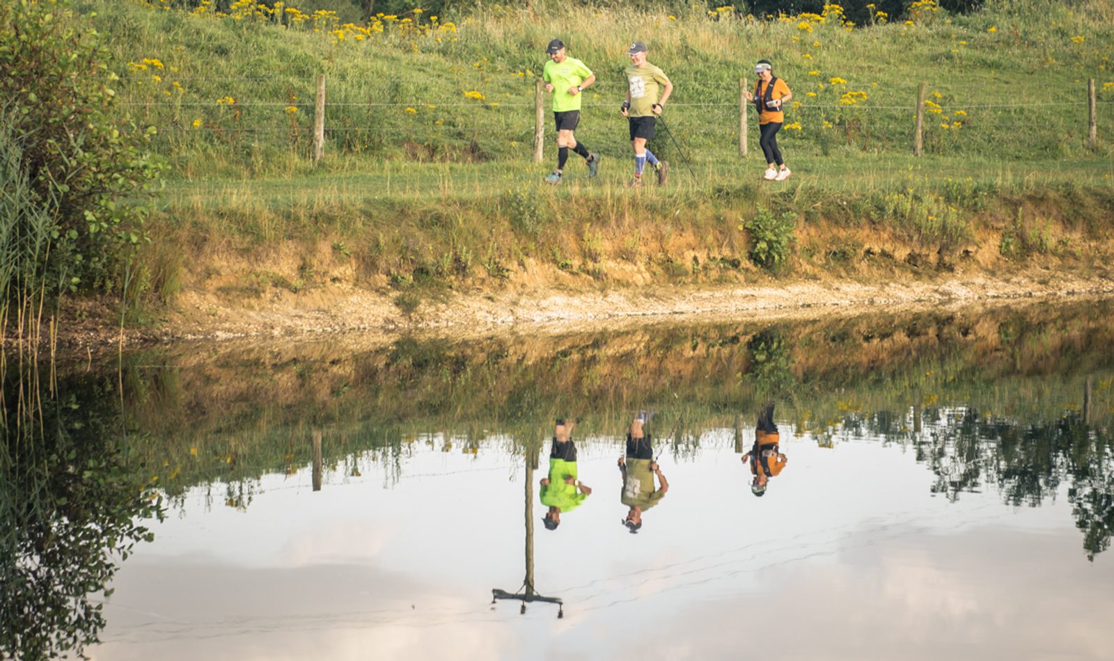 Four people are running along a grassy path next to a calm body of water. The water reflects the runners and the greenery around them. Yellow flowers dot the meadow, and wooden fence posts line the path. The sky is partly cloudy.