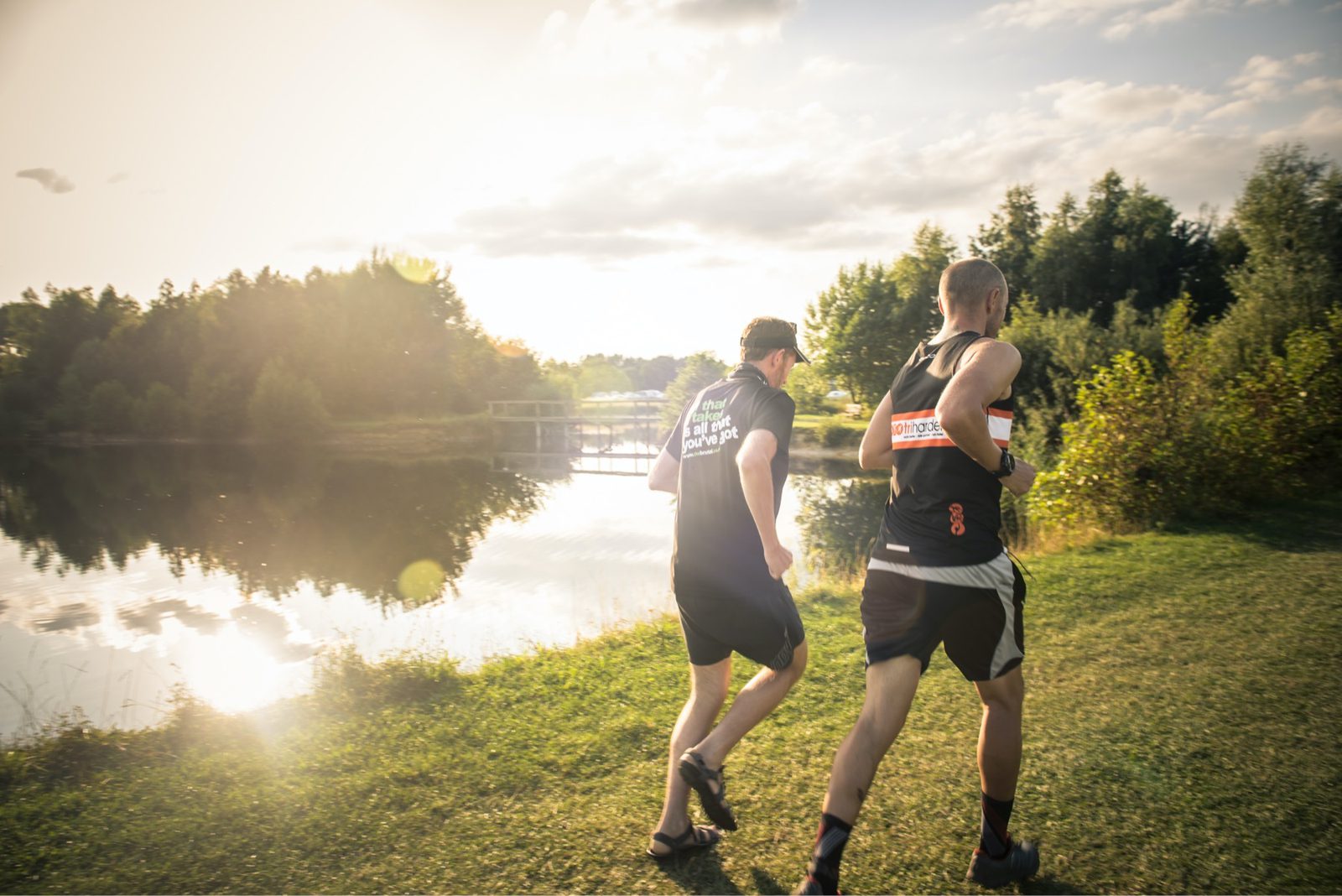 Two people are running on a grassy path beside a serene lake with reflections of trees and a small pier. They are dressed in athletic gear, and the sun is setting, casting a warm glow over the scene. Lush greenery surrounds the area.