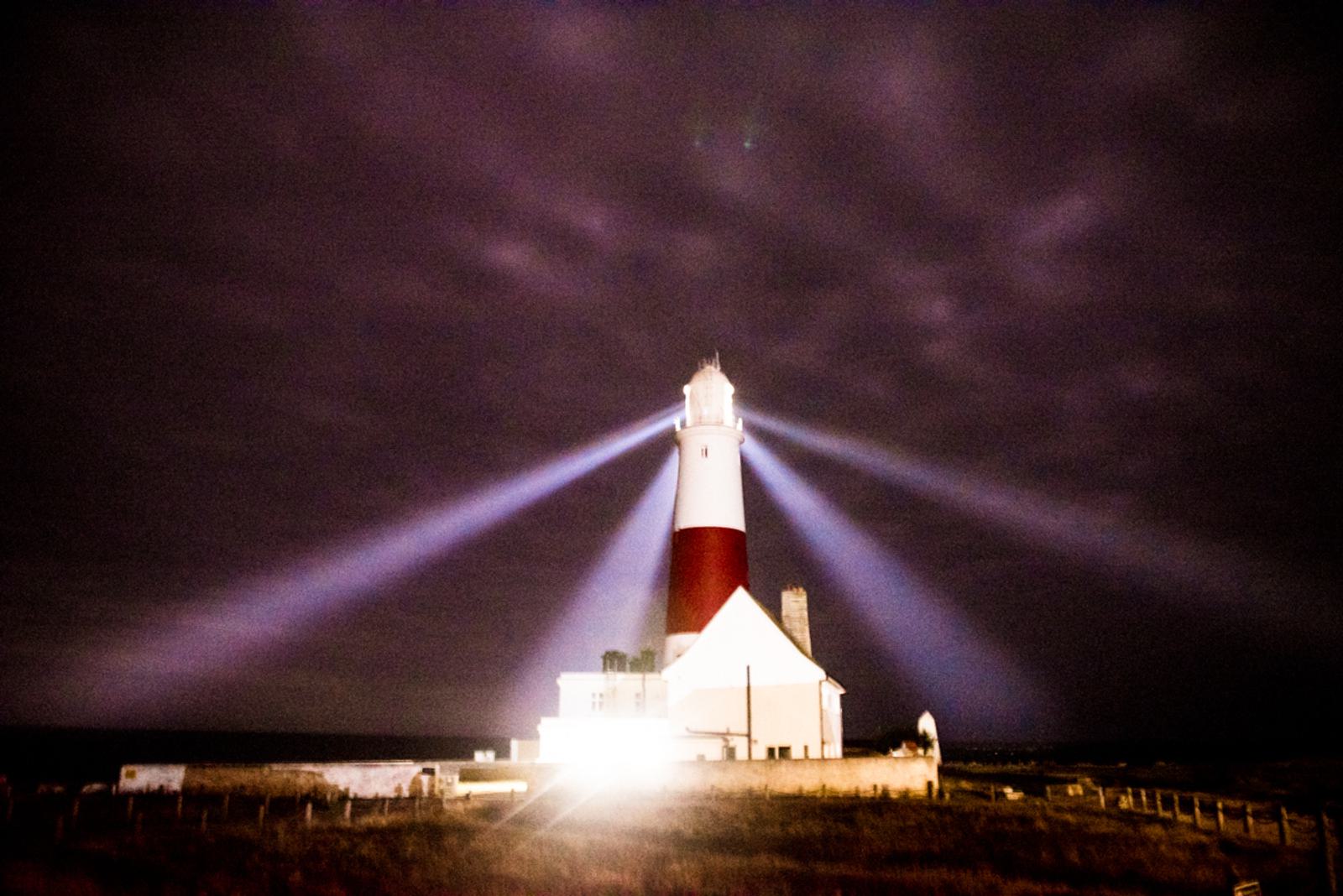 A lighthouse with a red and white striped tower shines multiple strong beams of light in different directions against a dark, cloudy night sky. The area around the lighthouse is dimly lit, highlighting its structure.