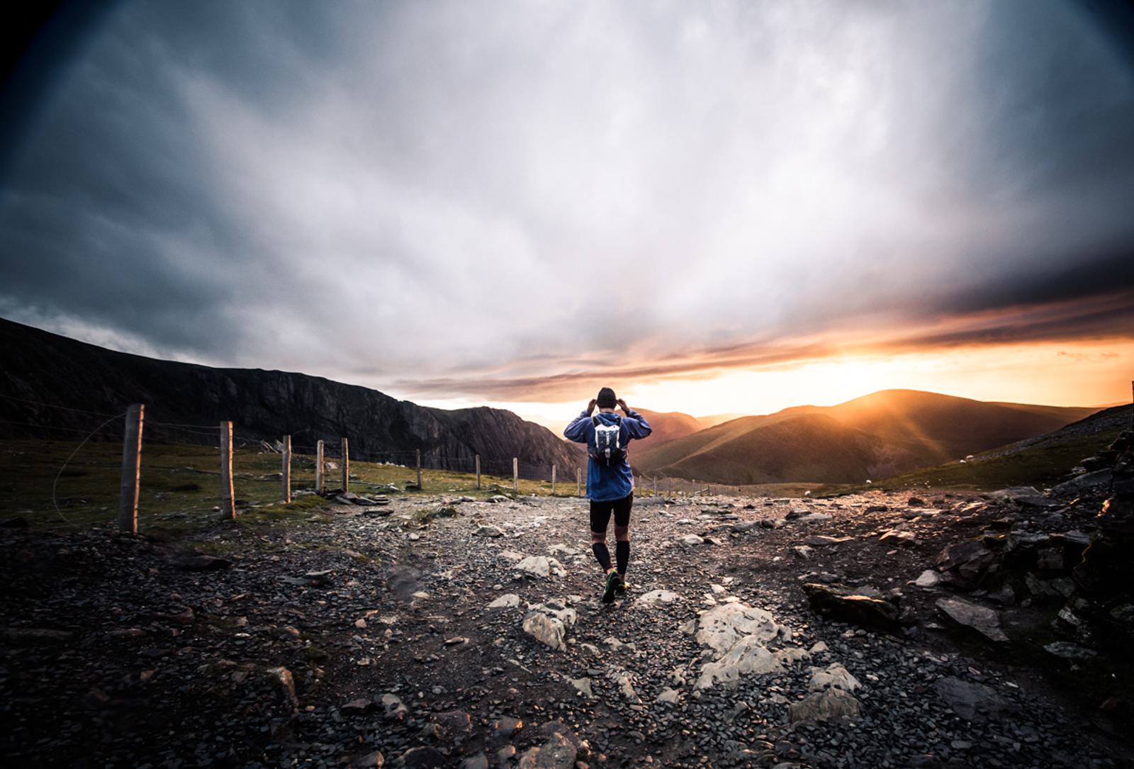 A person wearing a backpack walks on a rocky path towards a sunset, surrounded by mountains and a dramatic cloudy sky. The scene captures the serene beauty of nature with the horizon glowing in warm shades of orange and yellow.