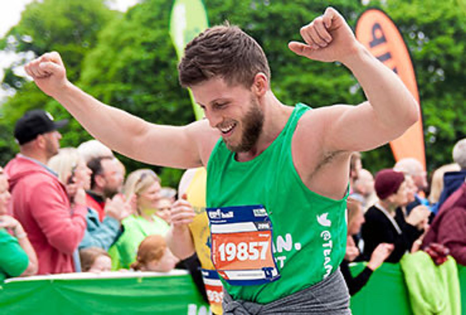 A runner, wearing a green tank top and bib number 19857, rejoices with both arms raised at the finish line of a race. A crowd of spectators claps and cheers in the background. Bright greenery is visible behind the crowd.