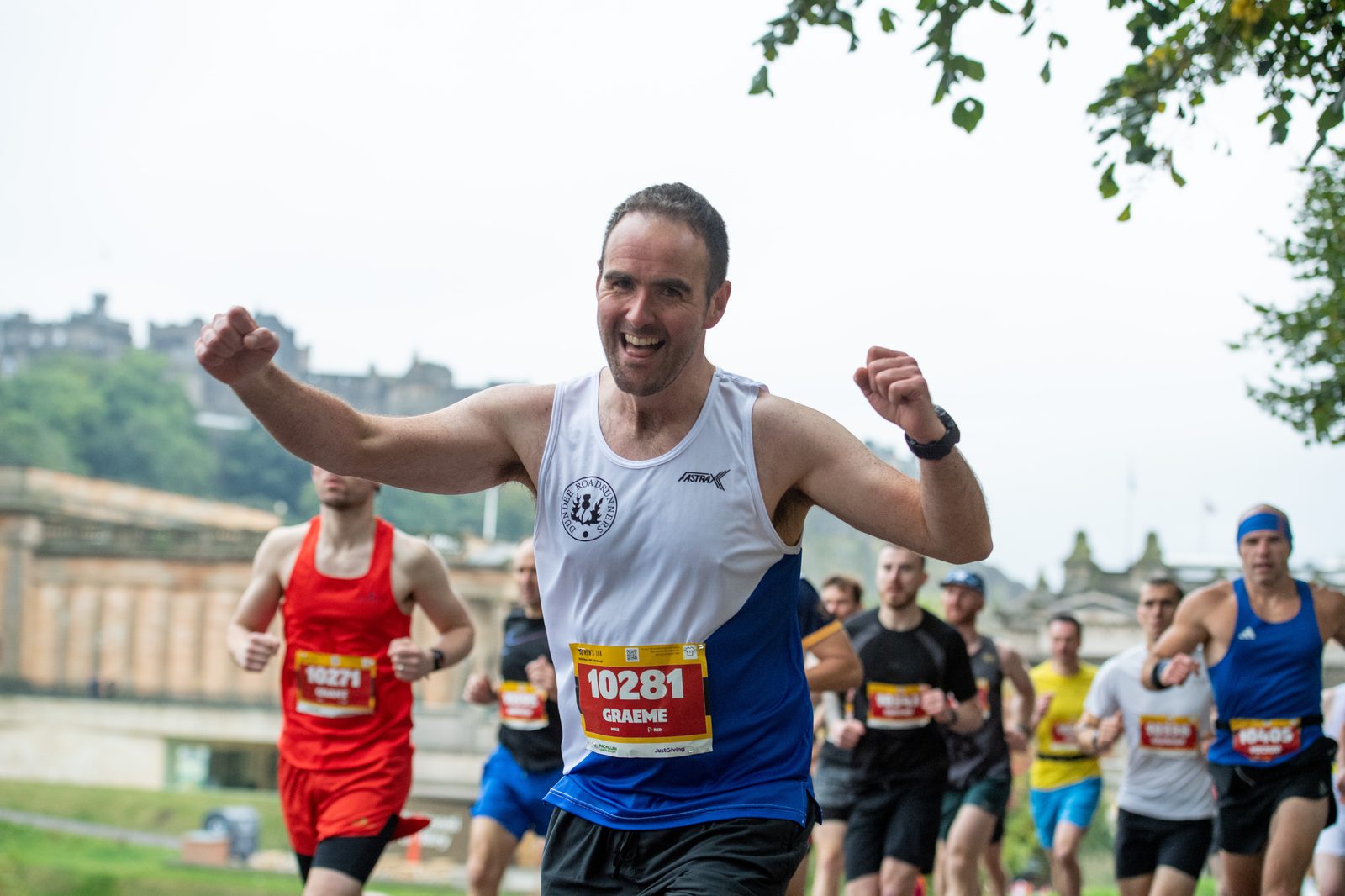 A group of runners participating in a race, with the focus on a smiling man in a white and blue tank top raising his arms in celebration. Other runners can be seen in the background, and trees partially frame the scene.
