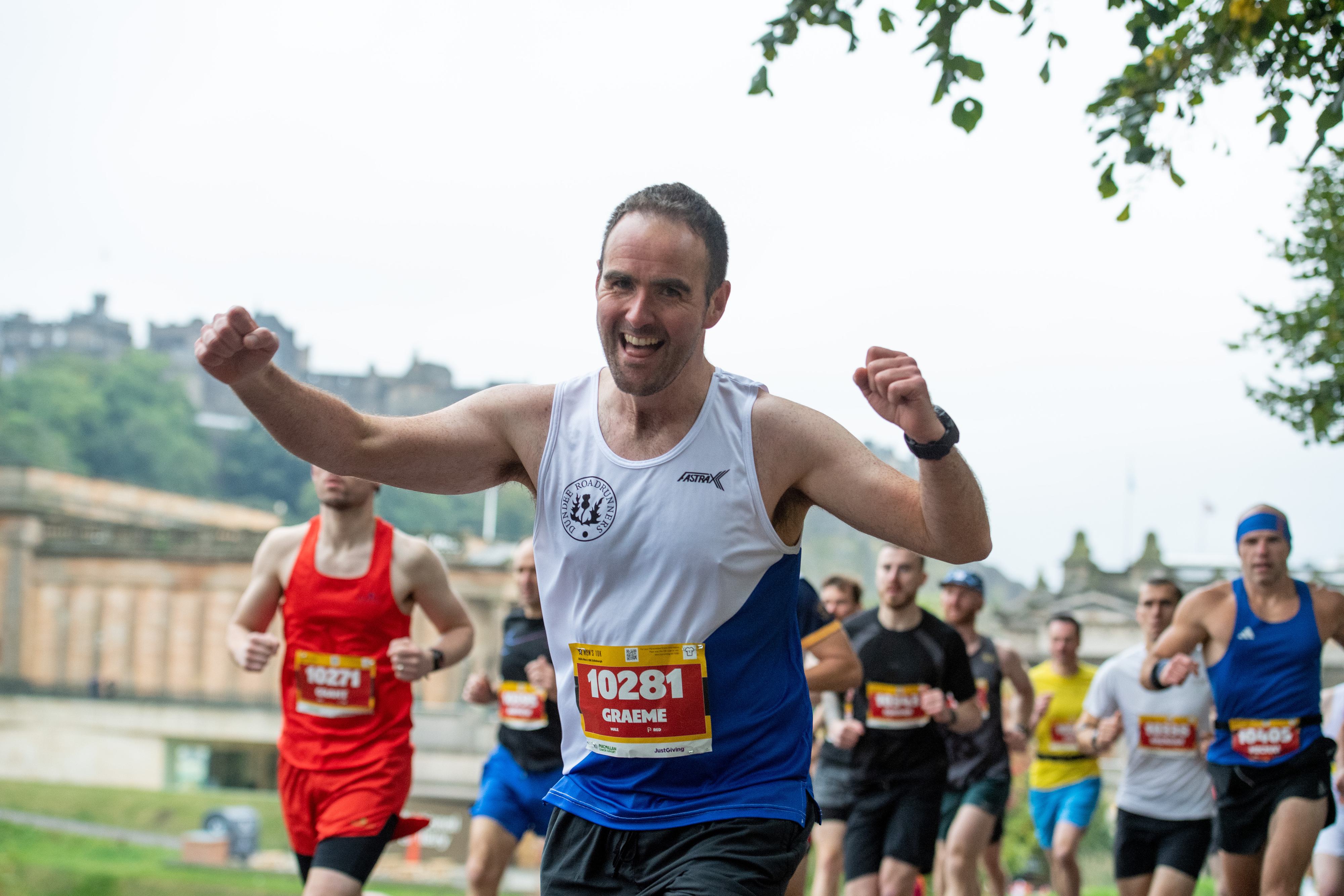 A group of runners participating in a race, with the focus on a smiling man in a white and blue tank top raising his arms in celebration. Other runners can be seen in the background, and trees partially frame the scene.