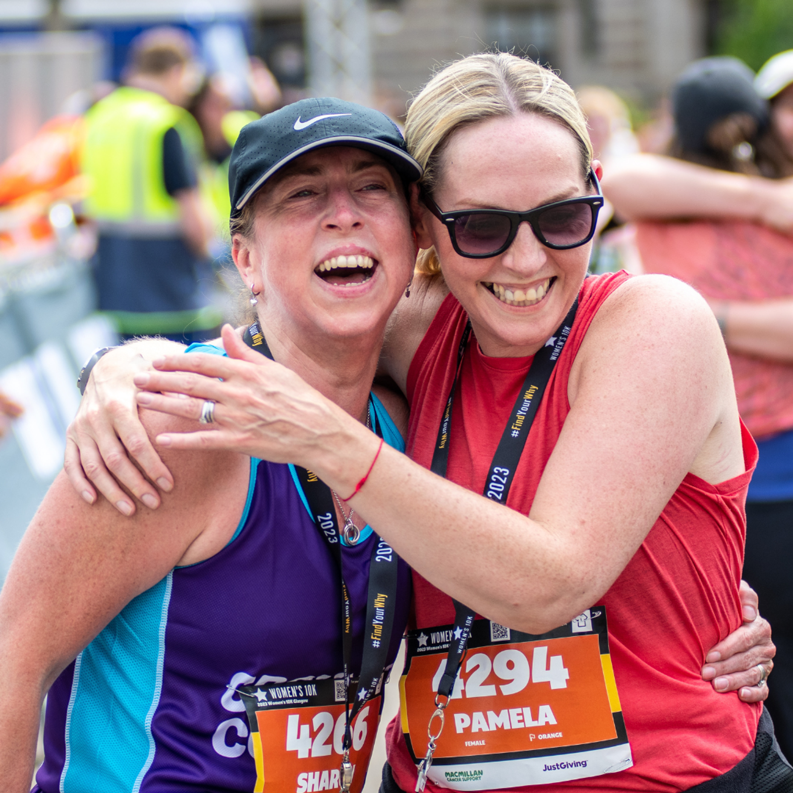 Two women are smiling and hugging after completing a race. They are wearing running bibs, medals, and athletic gear. The woman on the left is in a purple tank top and cap, while the woman on the right is in a red tank top and sunglasses.