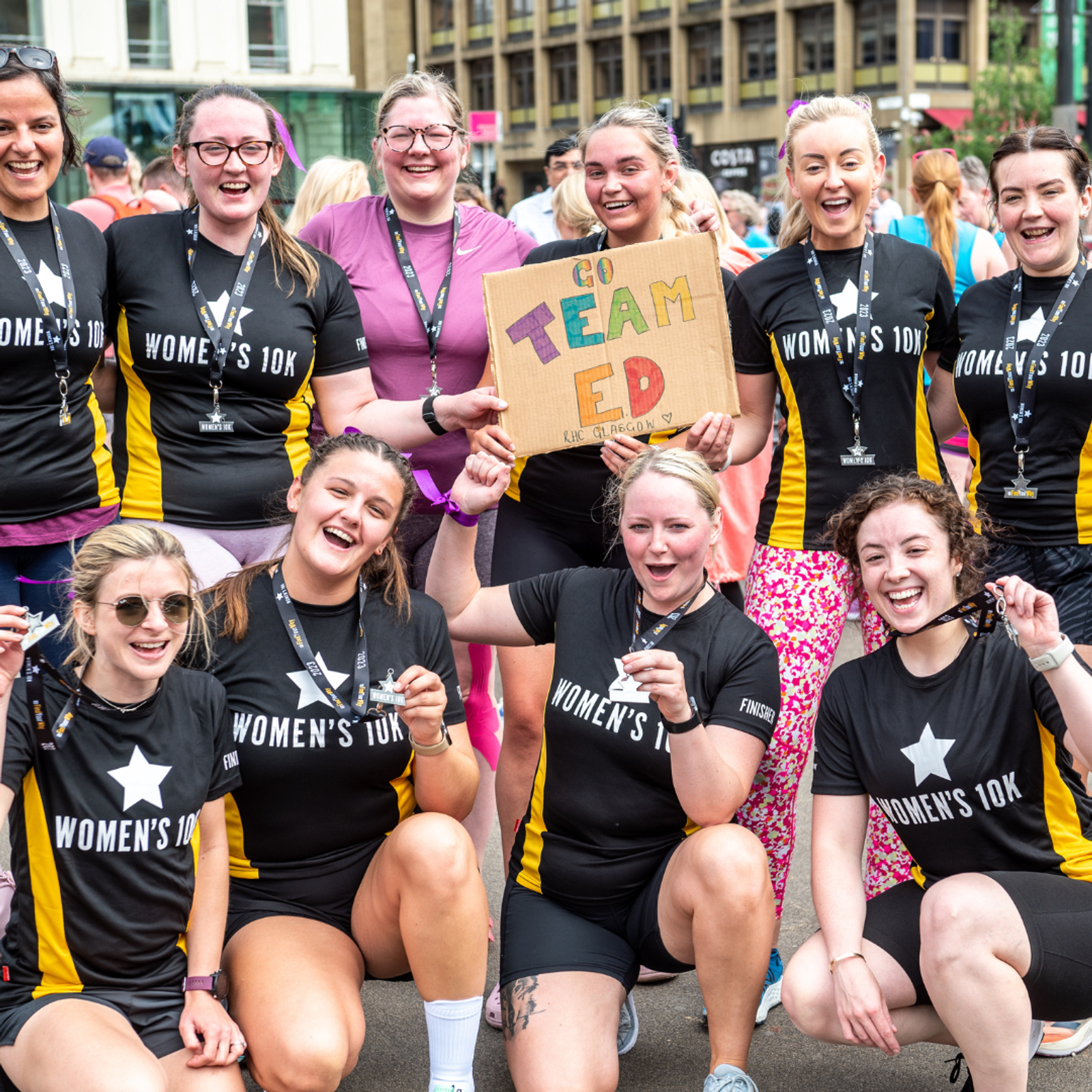 A group of women athletes, wearing black and yellow "Women's 10k" shirts, pose and smile together after a race. They hold medals and a cardboard sign that says "Go Team Ed!" in bright colors. The background shows other participants and buildings.