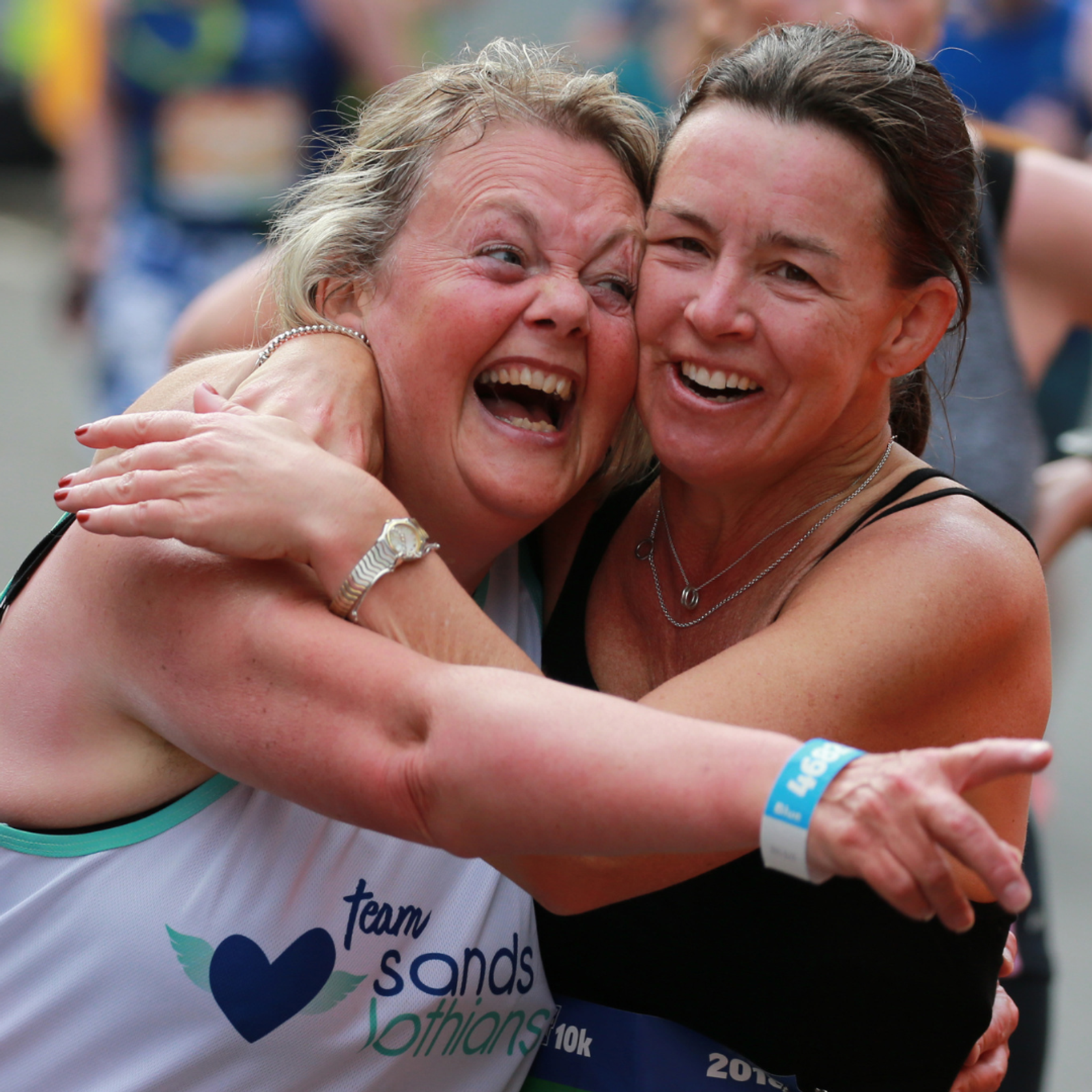 Two women, smiling broadly, embrace each other joyfully after completing a race. One wears a white shirt with the logo "team sands lothians," while the other sports a black athletic top. Both display exhaustion mixed with triumph, surrounded by fellow runners in the background.