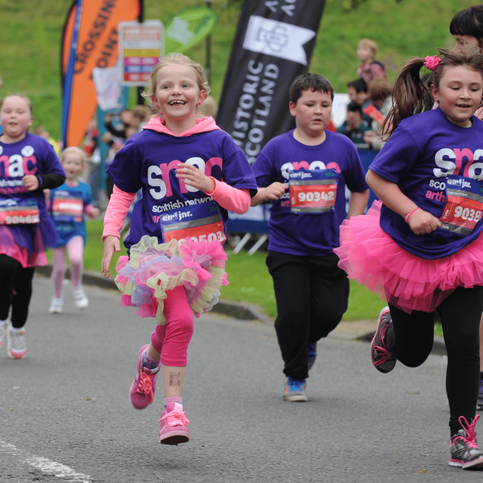 Young children in colorful tutus and purple shirts run joyfully during an outdoor race event, with spectators and banners visible in the background.