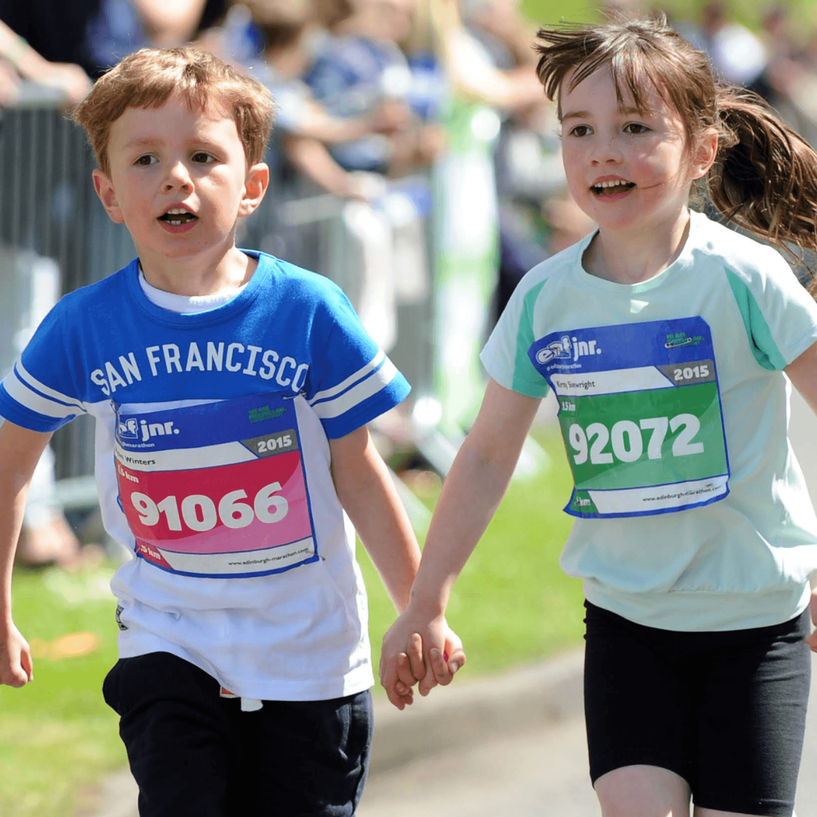Two young children holding hands and running in a race, wearing numbered race bibs and athletic clothing, with blurred spectators and barriers in the background on a sunny day.