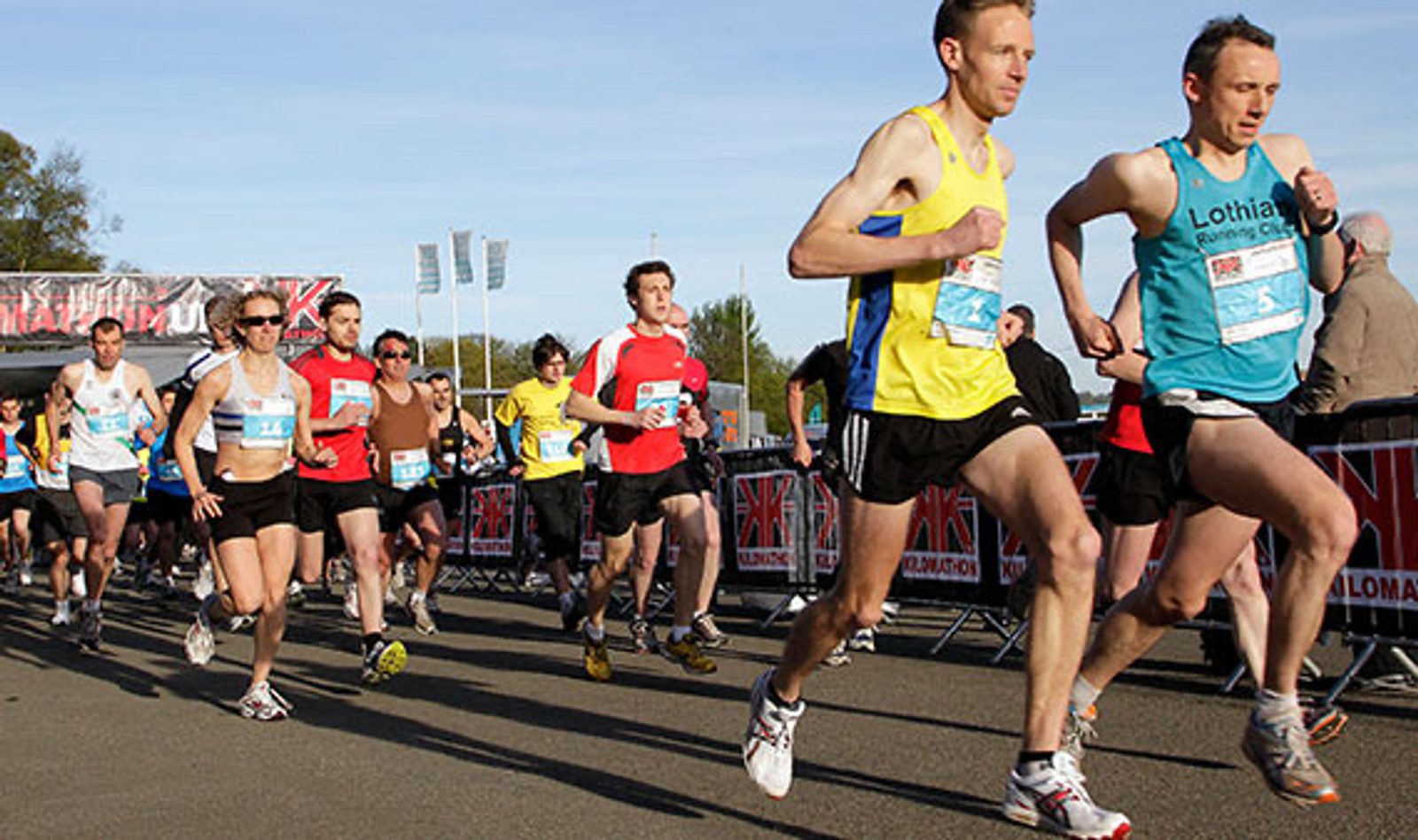 A group of runners, wearing race bibs and athletic gear, participate in a road race. Two runners in yellow and blue shirts are prominent in the foreground. Spectators and event banners can be seen in the background on a clear day.