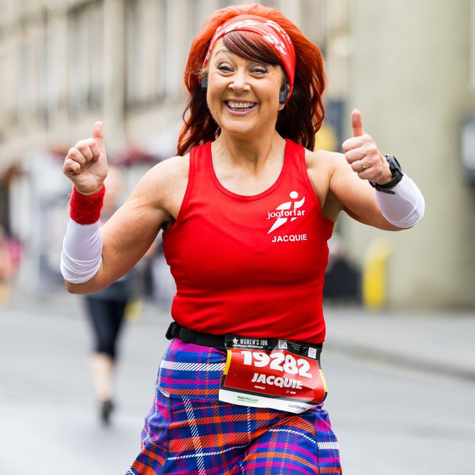 A woman wearing a red tank top and checkered skirt runs a race, smiling and giving thumbs up. She has a headband and a race bib with the number 19282. The background shows a blurred street scene.