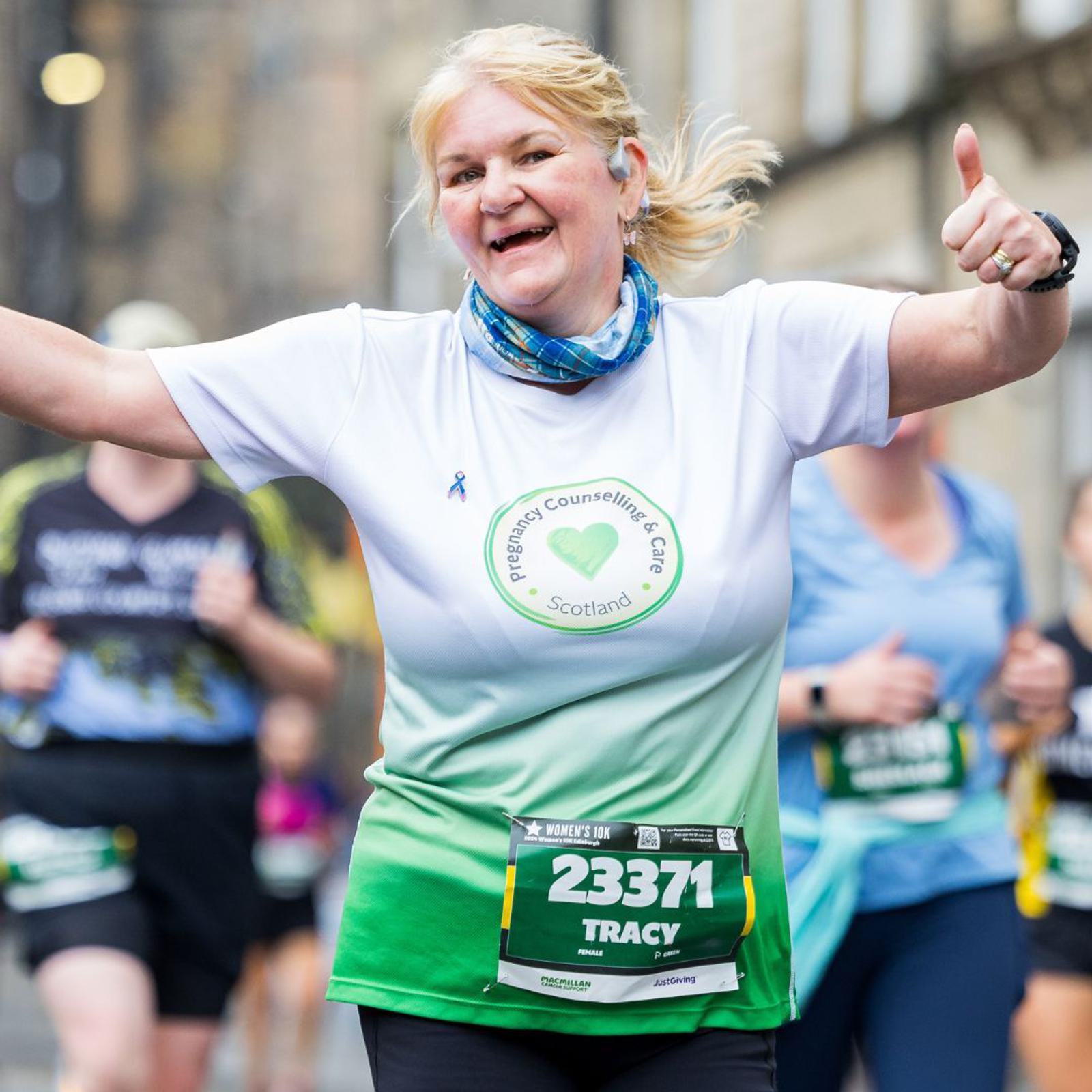 A woman is running in a marathon wearing a white and green shirt with "Pregnancy Counselling & Care Scotland" printed on it. She is smiling, with both arms raised, and wearing a race number 23371 labeled "Tracy." Other runners are in the background.