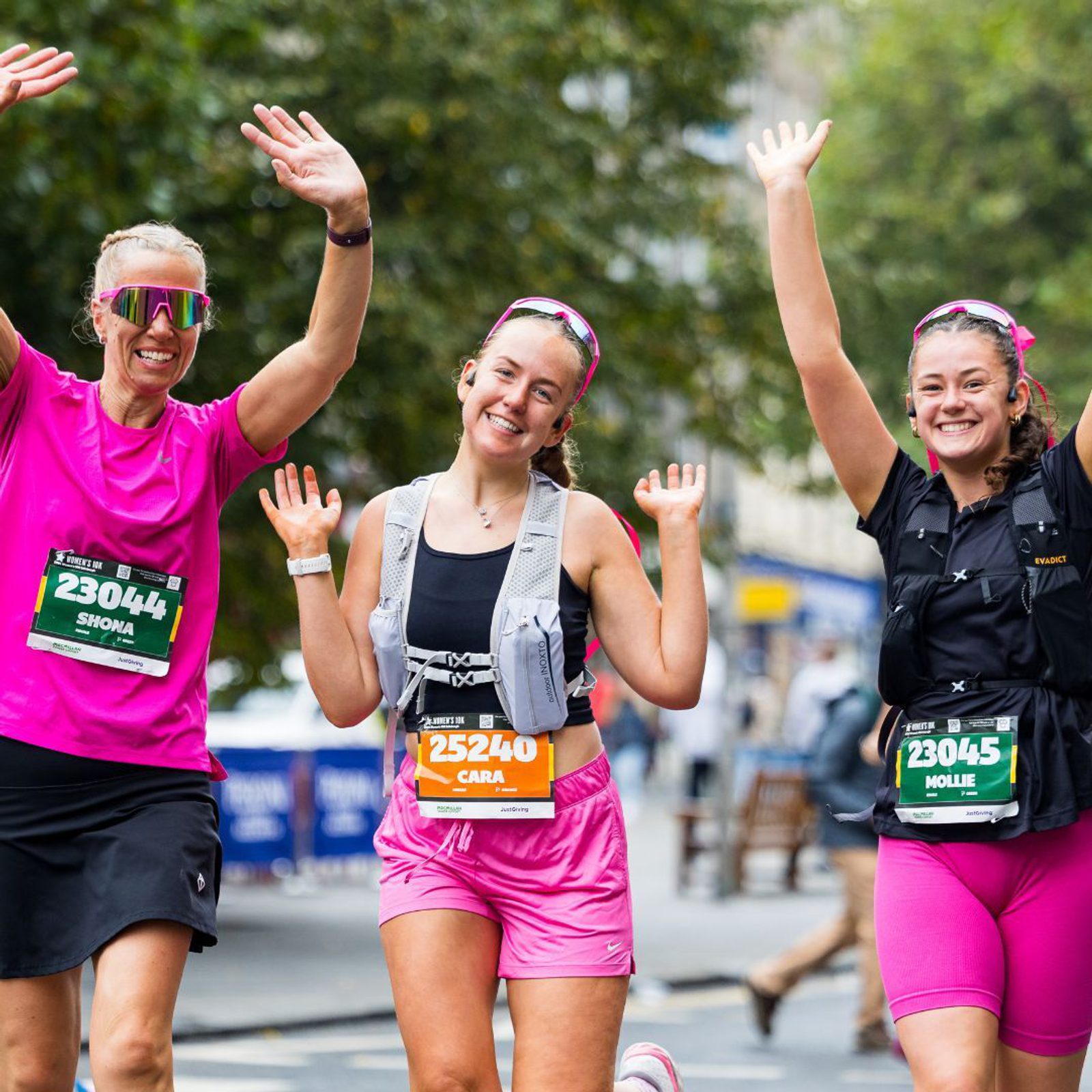 Three women in athletic gear are running, smiling, and waving at a marathon event. They are wearing colorful bibs with names and numbers. In the background, there are trees and blurred images of spectators.