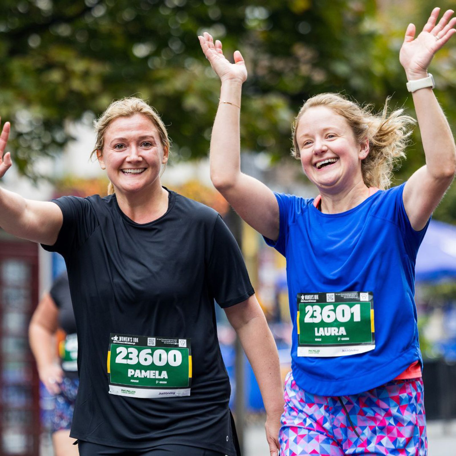 Two women in athletic wear, wearing race bibs numbered 23600 and 23601, smile and raise their arms as they participate in a running event. Trees and other runners are visible in the background.