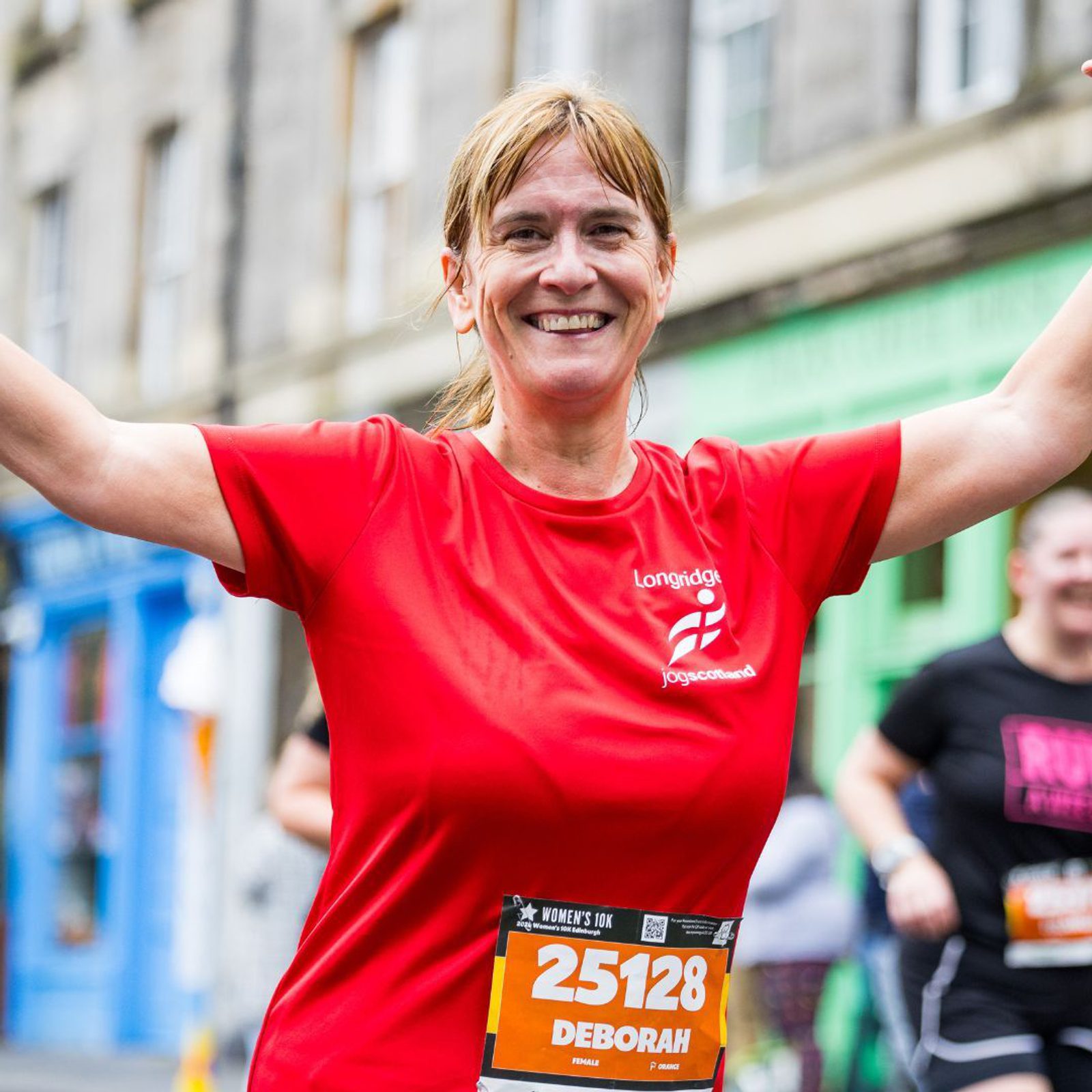 A smiling woman wearing a red t-shirt and race bib number 25128 runs with her arms raised. The background shows a street with colorful buildings and other runners.