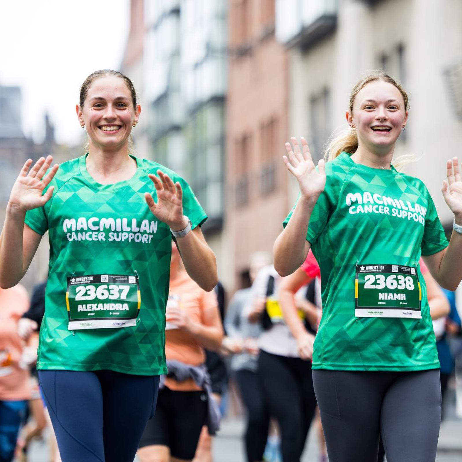 Two women in green Macmillan Cancer Support shirts wave at the camera while running in a marathon. They are wearing bib numbers 23637 and 23638. A crowd of runners and blurred urban buildings are in the background.