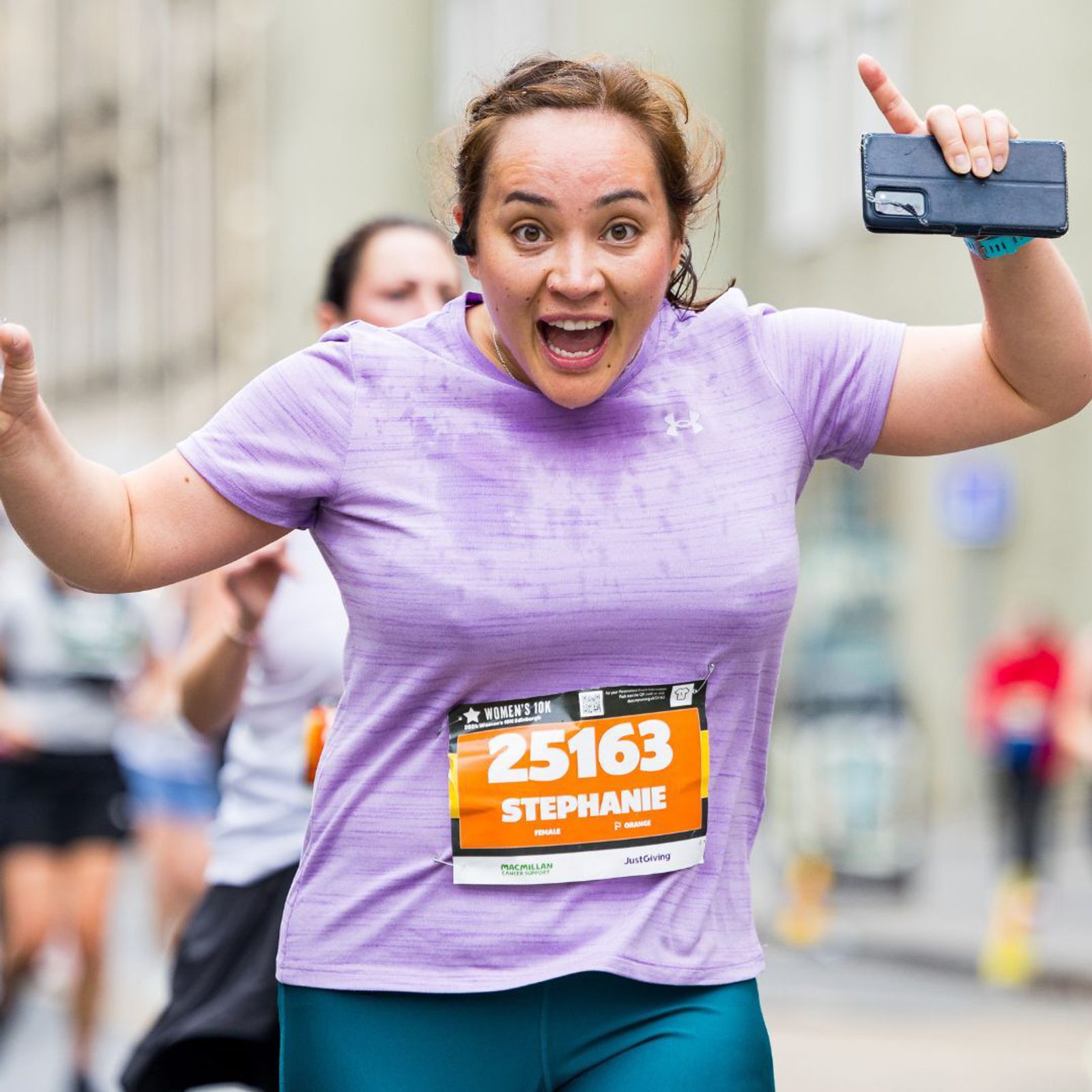 A woman in a light purple shirt and turquoise pants is running in a race, smiling joyfully with her arms raised. She has a race bib with the number 25163. In her hand, she's holding a smartphone. Other runners are visible in the background.
