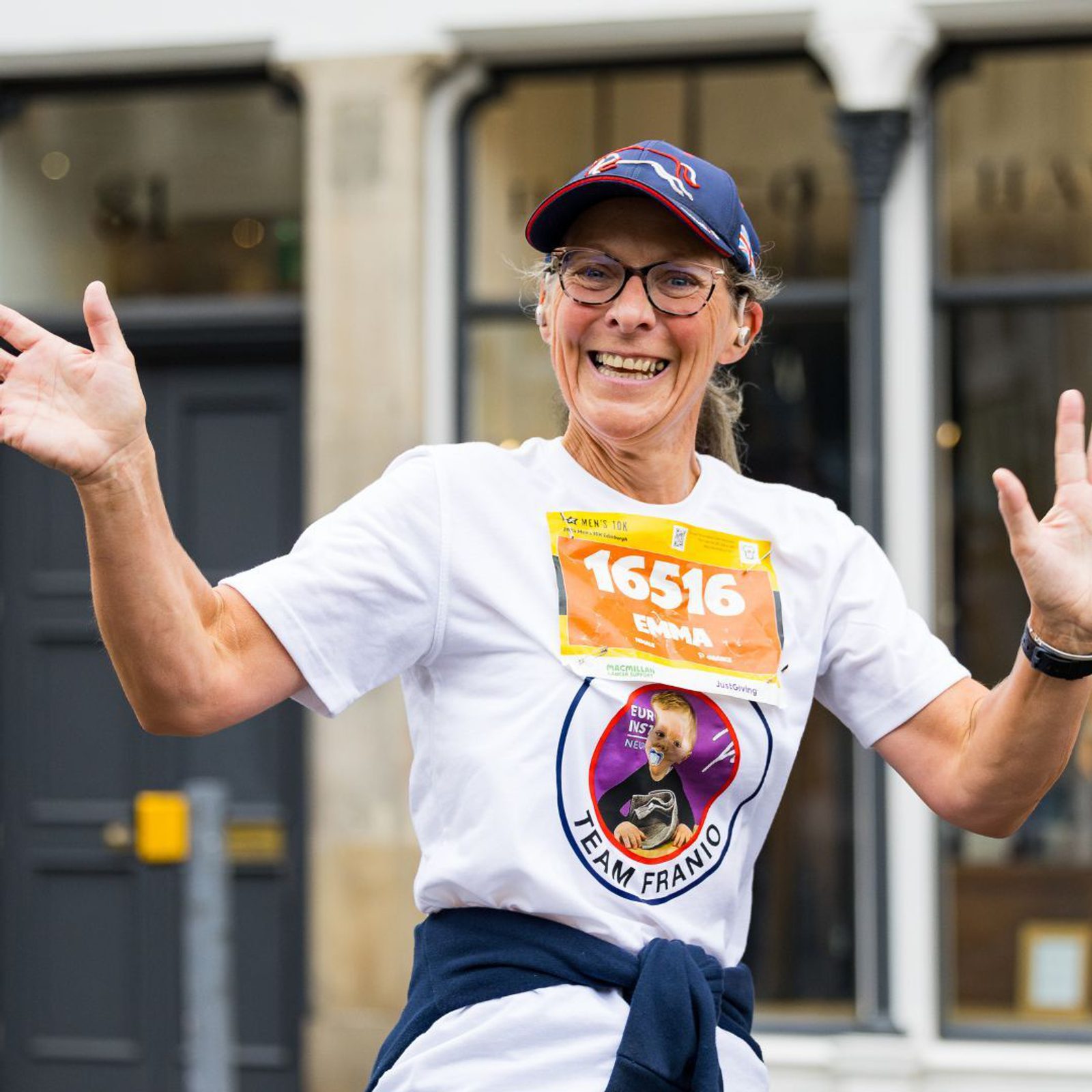 A person wearing glasses and a cap smiles and waves while running. They have a race bib with the number 16516. They're wearing a white "Team Franjo" shirt with a photo of a person on it, and have a dark sweater tied around their waist.