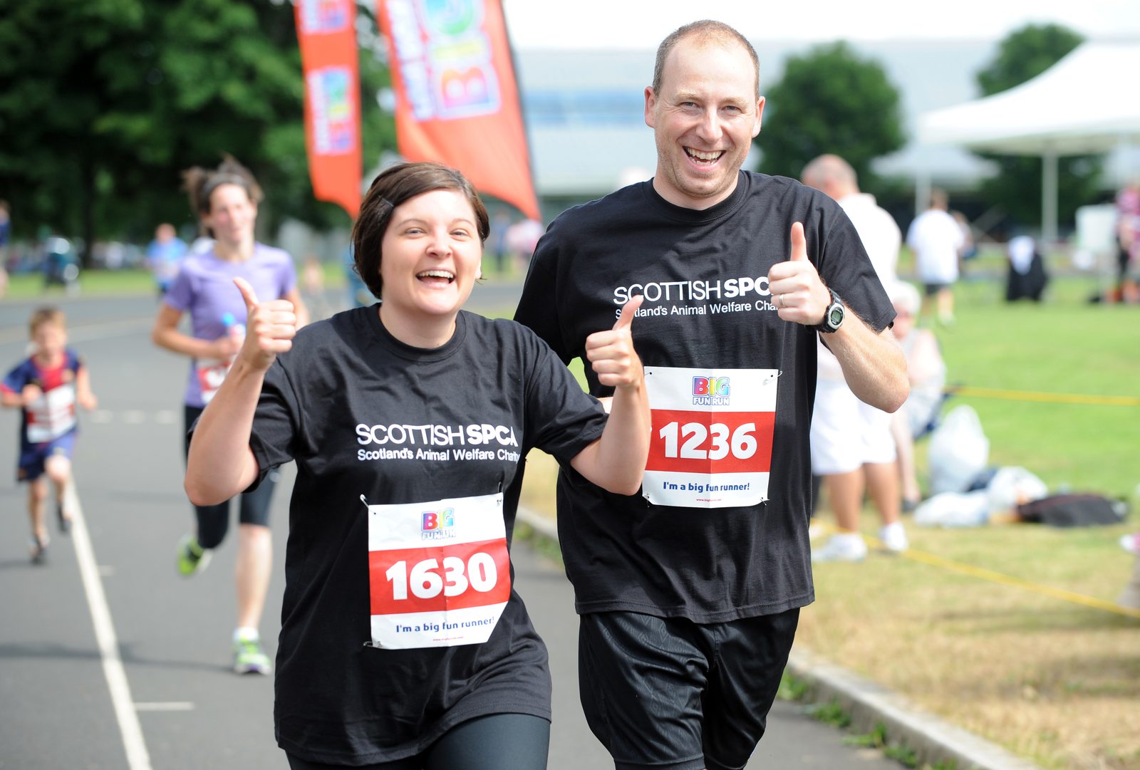 Two smiling runners, a woman and a man, wearing 'Scottish SPCA' T-shirts, run in an outdoor event. They show enthusiasm with thumbs-up gestures and display race bibs numbered 1630 and 1236. Other runners and event tents are visible in the background.