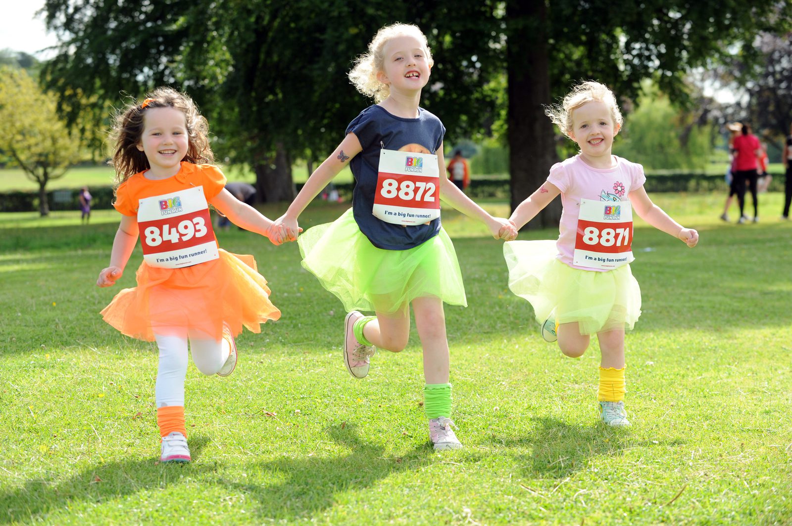 Three young children are running on grass during an outdoor event. They wear colorful tutus over shirts and shorts, each with a bib number: 8493, 8872, and 8871. They appear joyful and are holding hands. Trees and people are visible in the background.