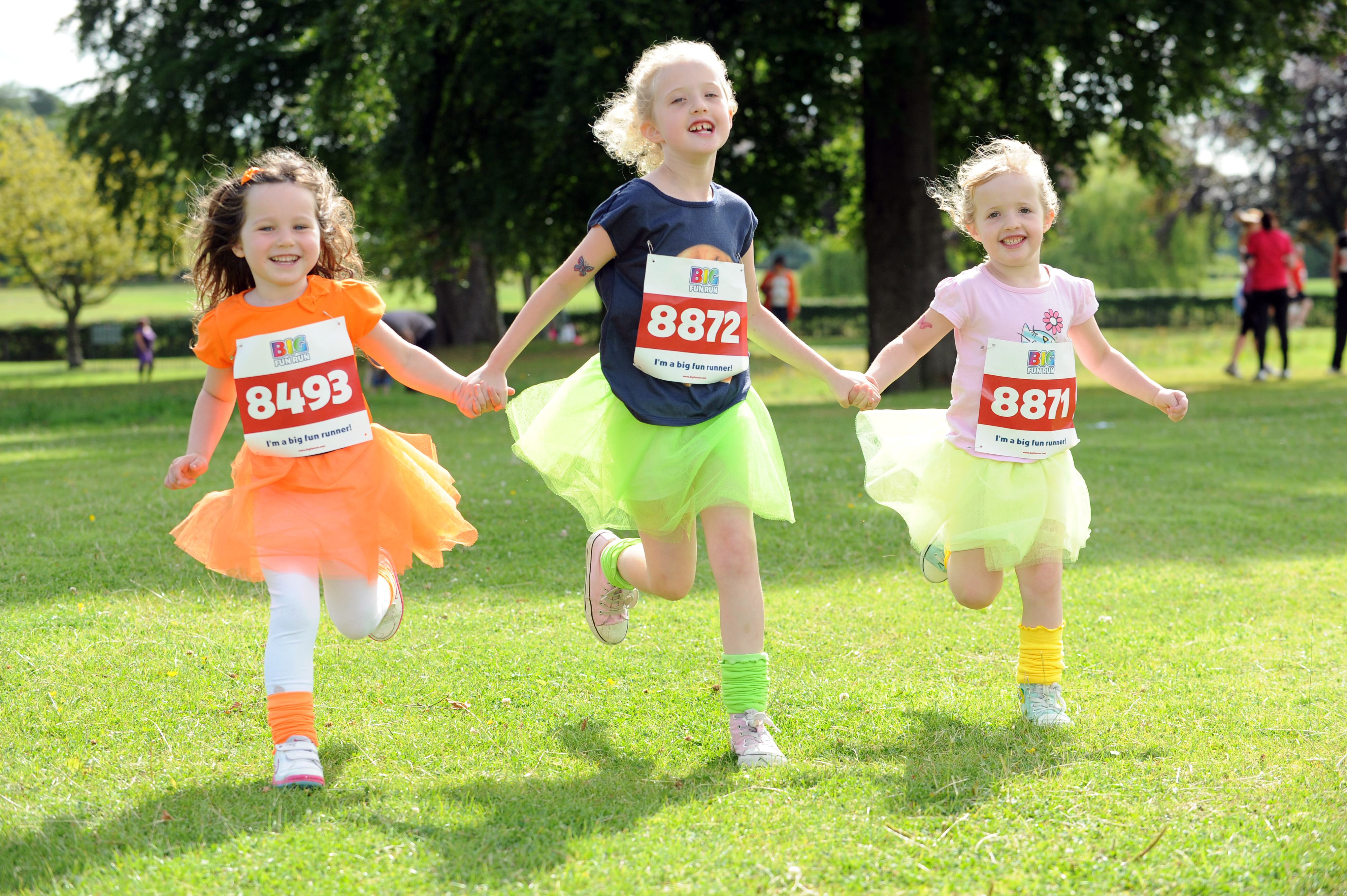Three young children are running on grass during an outdoor event. They wear colorful tutus over shirts and shorts, each with a bib number: 8493, 8872, and 8871. They appear joyful and are holding hands. Trees and people are visible in the background.