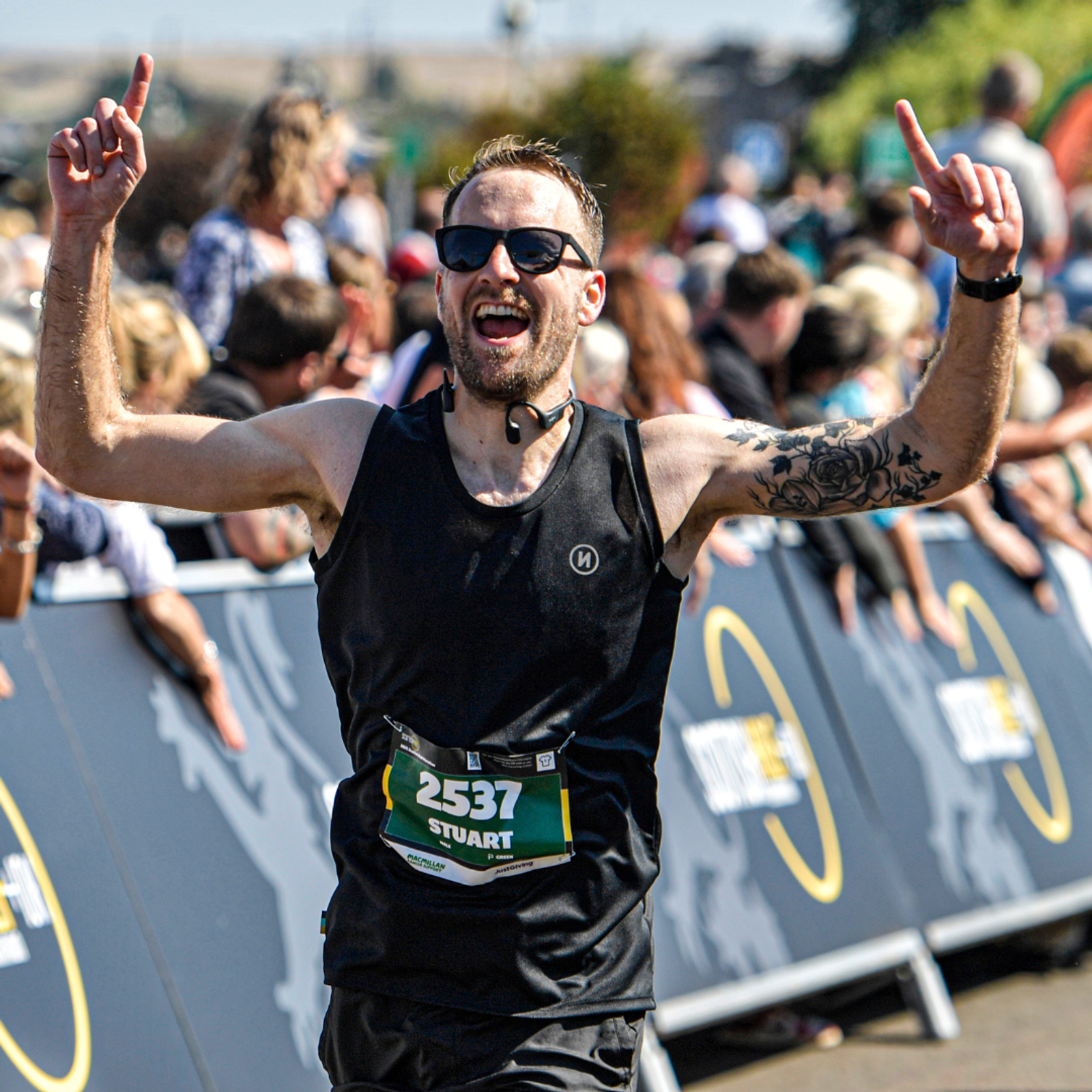 A male runner wearing sunglasses, a black tank top, and a race bib labeled "2537 STUART" celebrates with both arms raised at a crowded outdoor race finish line.