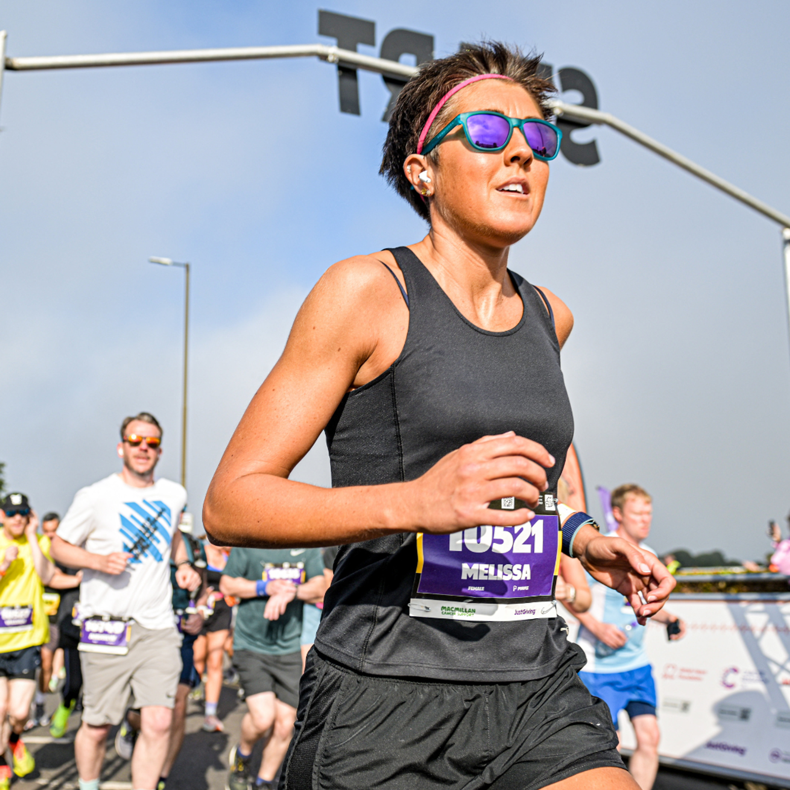 A woman wearing sunglasses, a black tank top, and a race bib runs in a road race, surrounded by other runners. The sky is partly cloudy and there is a large sign structure in the background.