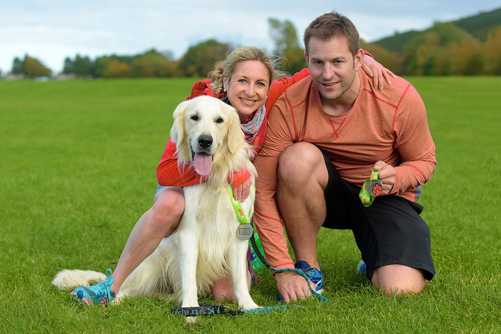 A woman and a man are kneeling on a grassy field with a Golden Retriever. The dog is wearing a medal around its neck, and both the woman and the man are holding medals. They are all smiling at the camera, with trees and hills in the background.