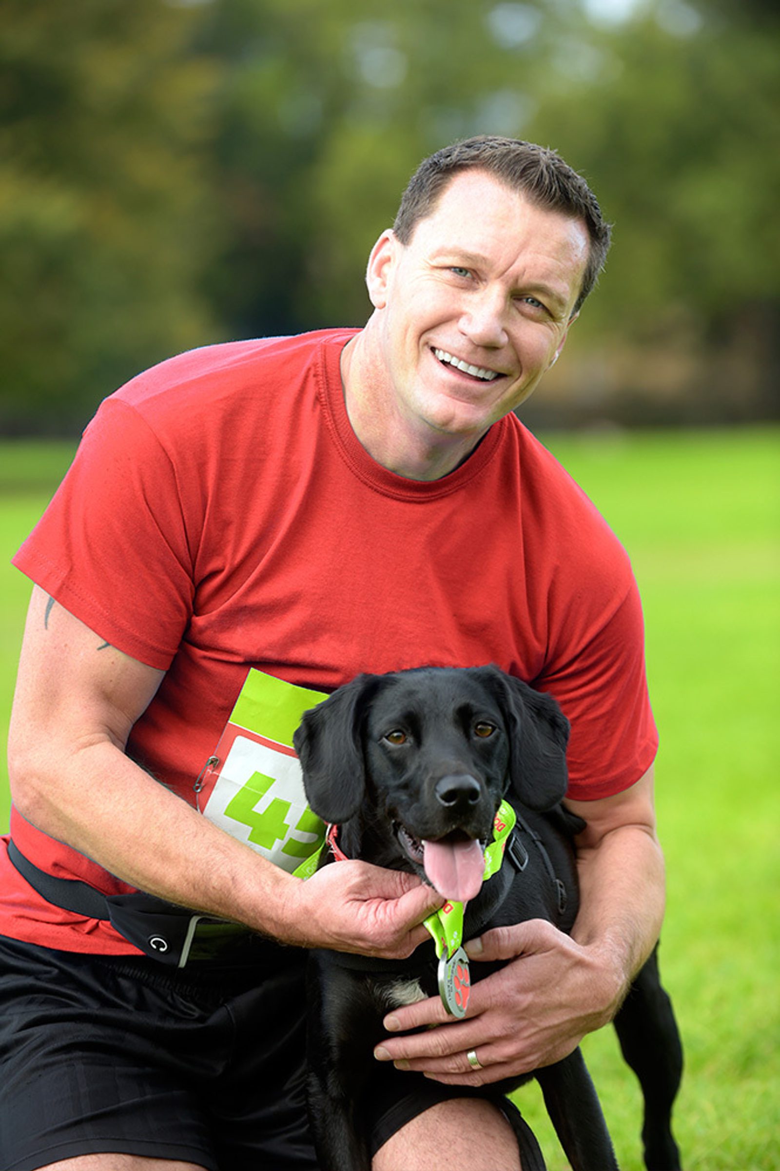 A man in a red T-shirt kneels on the grass, smiling broadly while holding a black Labrador wearing a medal. Both seem happy and energetic with a green scenic background. The man has an event number pinned to his shirt.