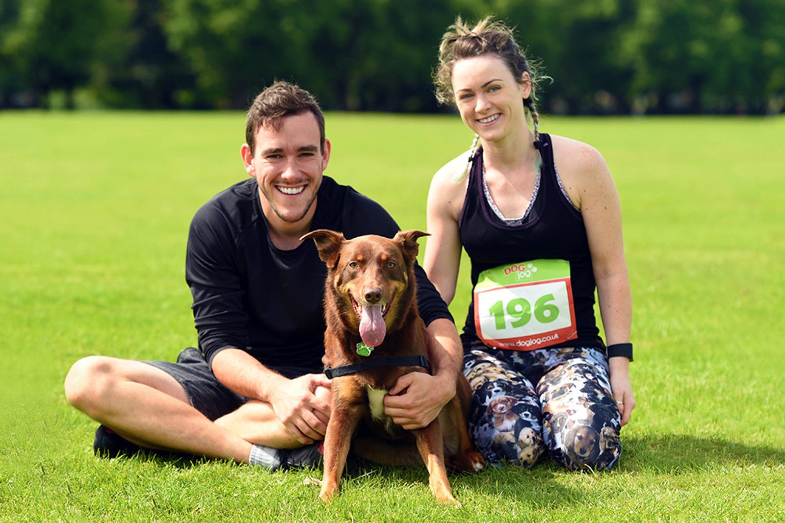 A man and a woman, both dressed in athletic clothing and race bibs, sit on the grass in a park with a happy brown dog between them. The woman is wearing bib number 196. They are smiling and appear relaxed, enjoying a sunny day outdoors.