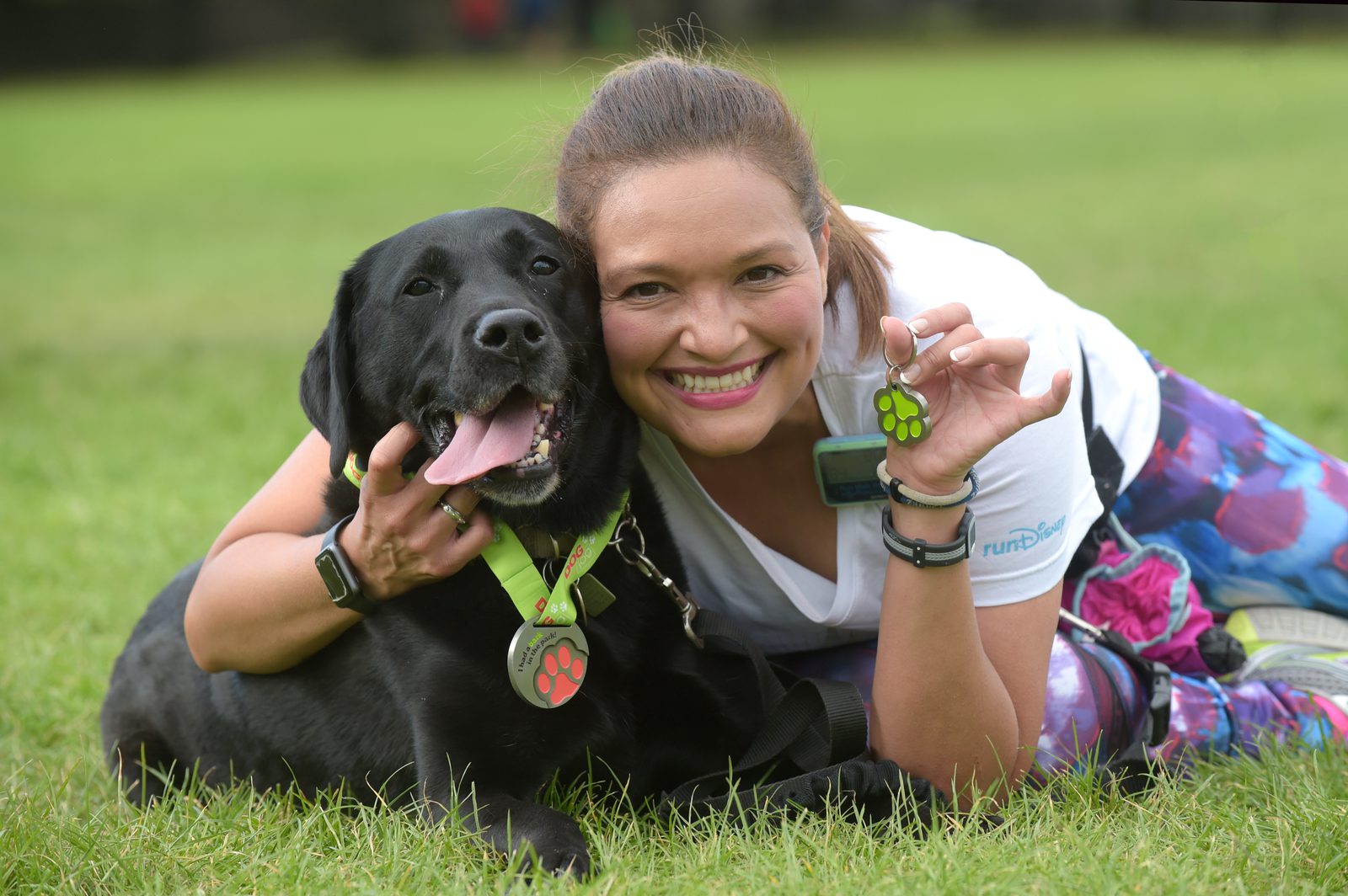 A woman lies on the grass, smiling and holding a dog tag with a green paw print. Beside her is a black Labrador retriever, also wearing a collar with a green paw print tag. They appear happy and content outdoors in a grassy field.