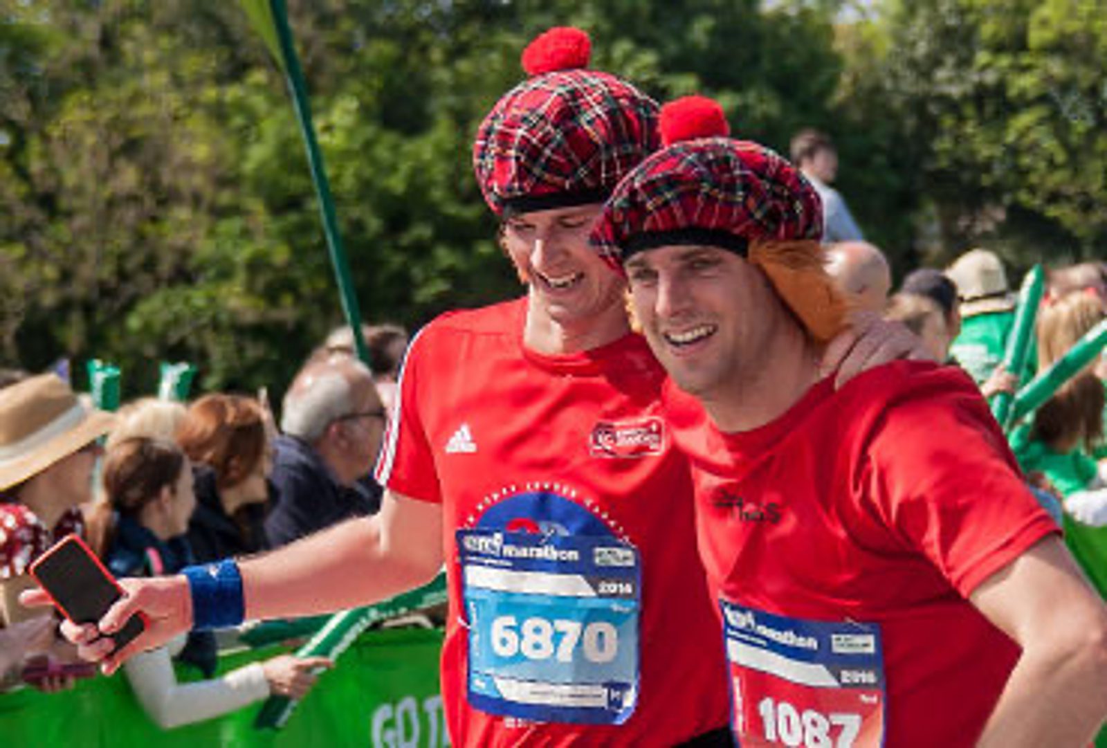 Two men in red T-shirts and checkered hats with red pom-poms, smiling and posing for a photo at a marathon event. One holds a smartphone, and both have race numbers pinned to their shirts. People and trees are visible in the background on a sunny day.