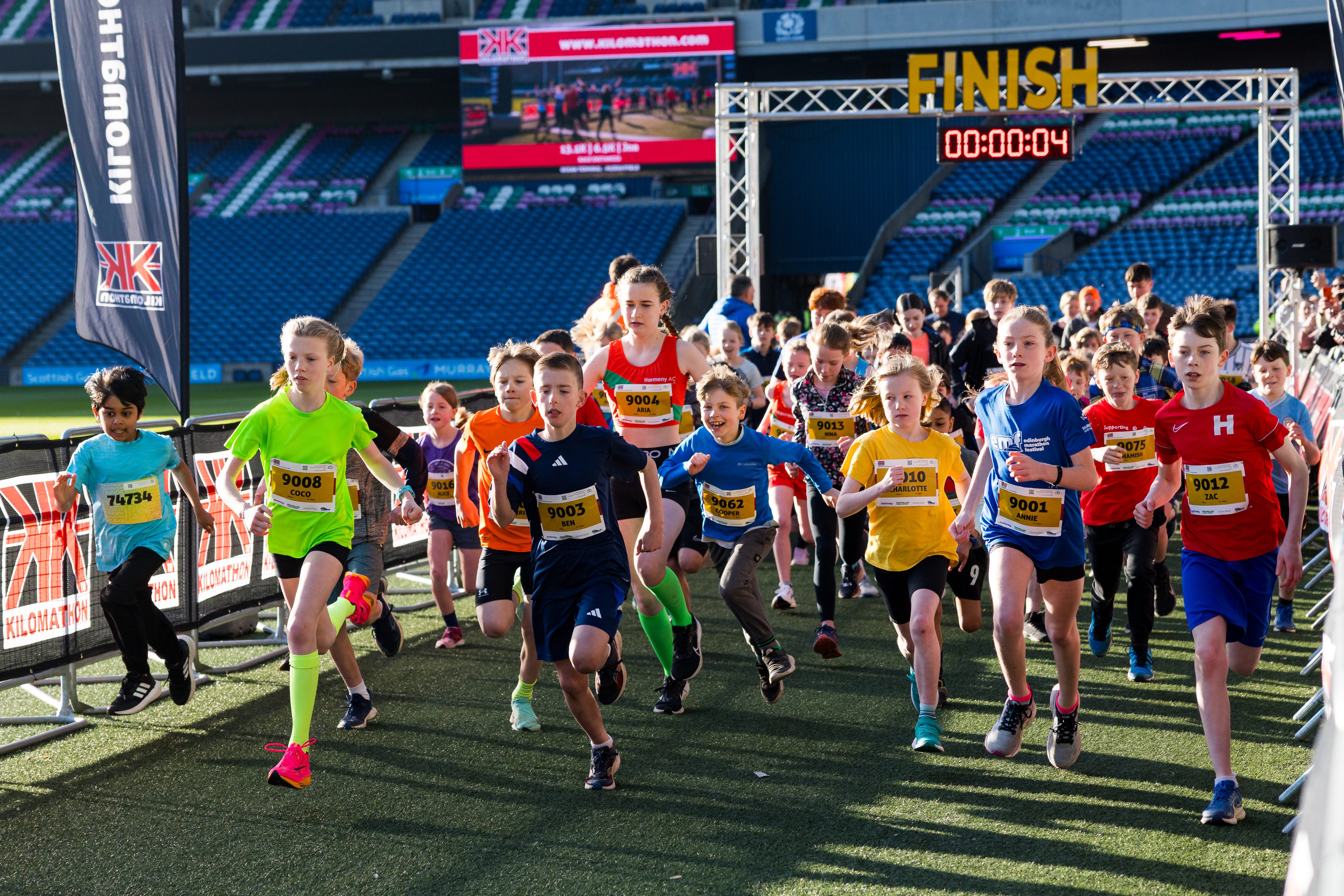 A group of children in colorful sports clothing starts running from a starting line at a race event in a stadium, with a large "FINISH" sign and digital timer overhead. Spectators and other participants watch in the background.