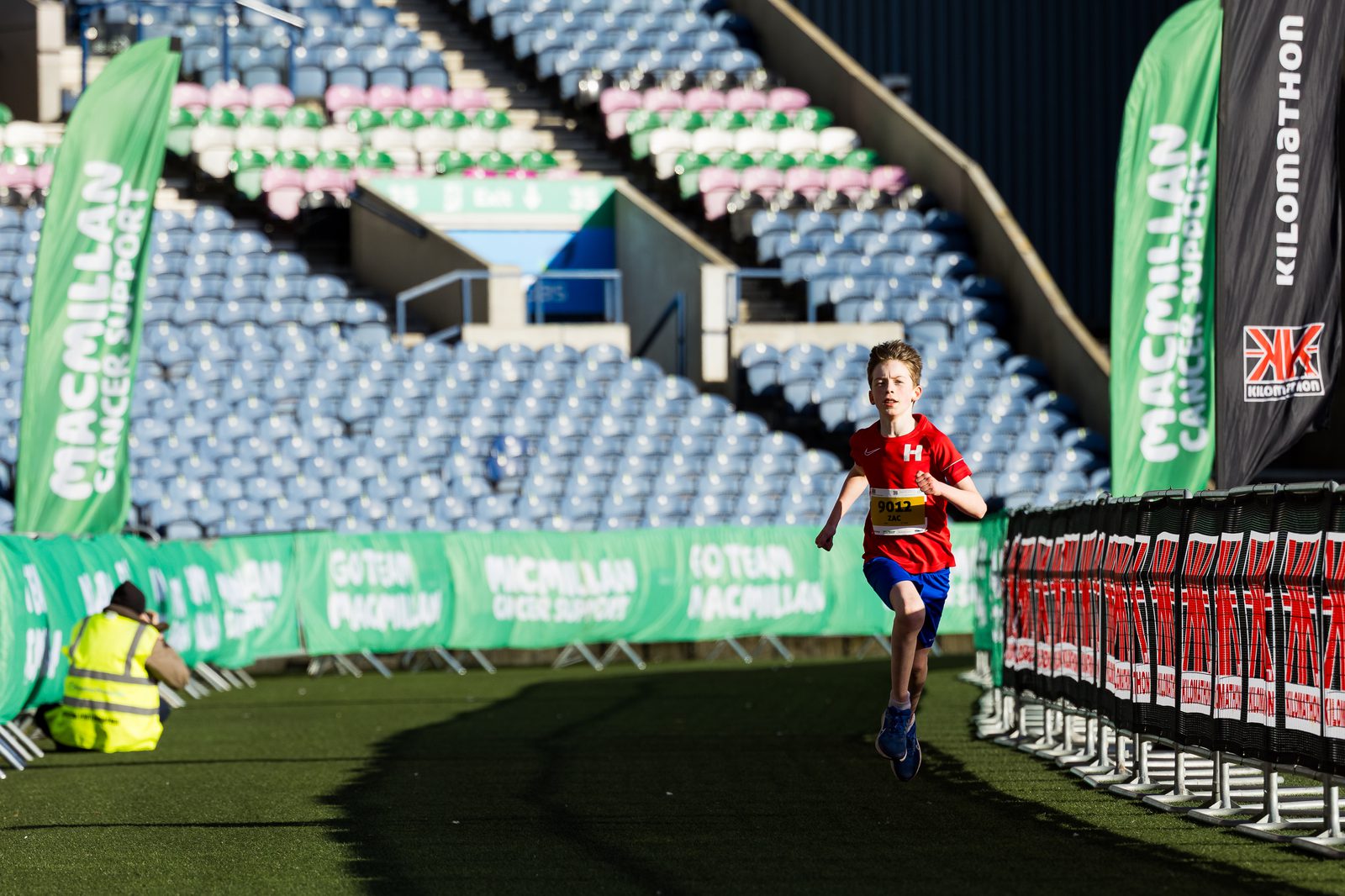 A young boy in a red shirt runs alone on a stadium track during a race, with empty seats in the background and green Macmillan Cancer Support banners along the route.