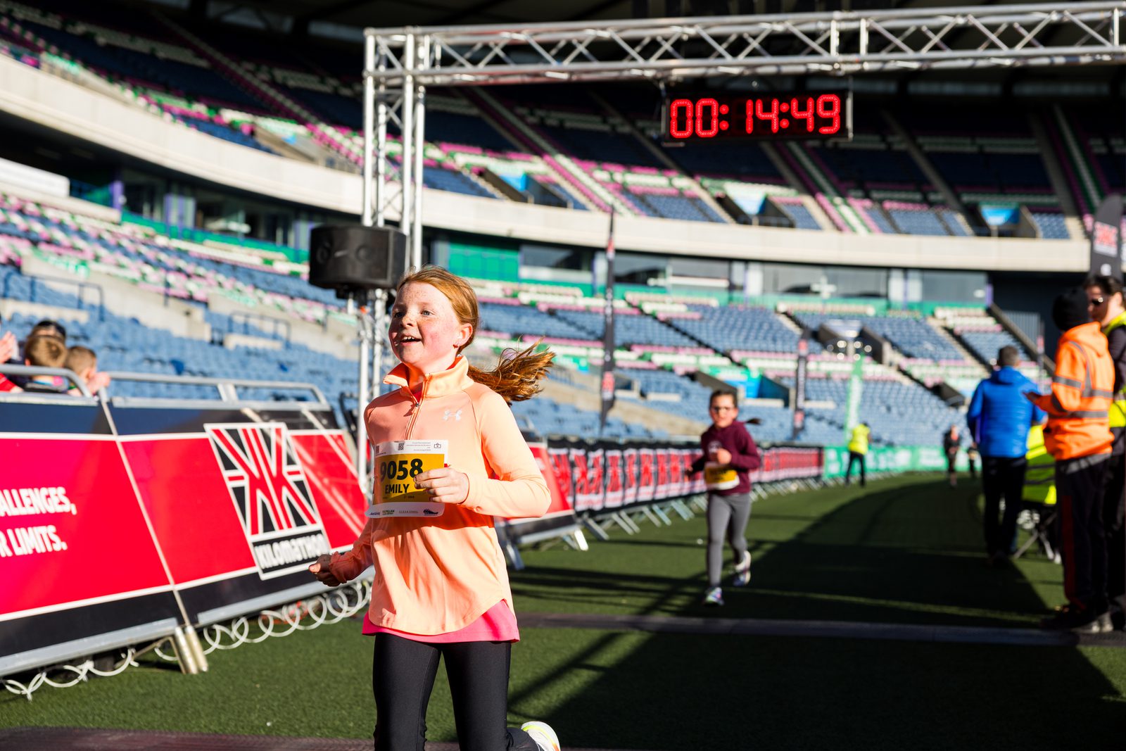 A young girl in a pink top runs toward the finish line at a stadium race, smiling with her race number visible. A digital timer above shows 14:49, and several people are visible in the background.