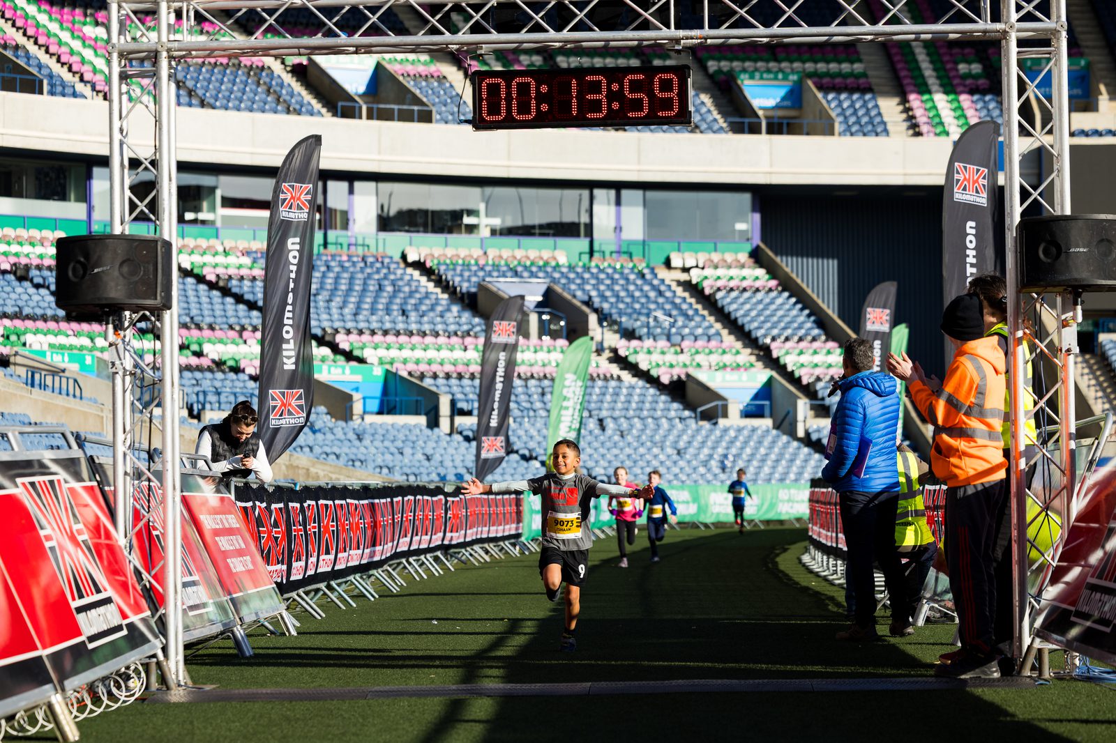 A young runner sprints toward the finish line of a race, arms outstretched, as the digital clock above shows 13:59. Spectators and staff in bright jackets watch from the sideline in a stadium.
