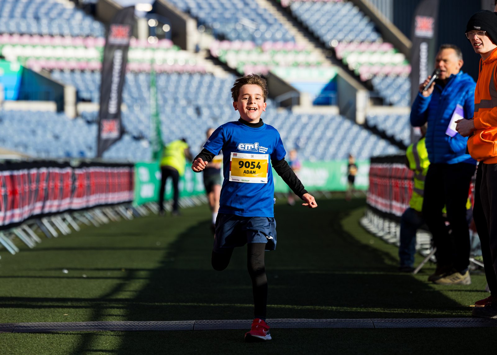 A young boy wearing a blue shirt and race bib number 9054 sprints joyfully toward the finish line at a stadium event, with spectators and officials in the background.