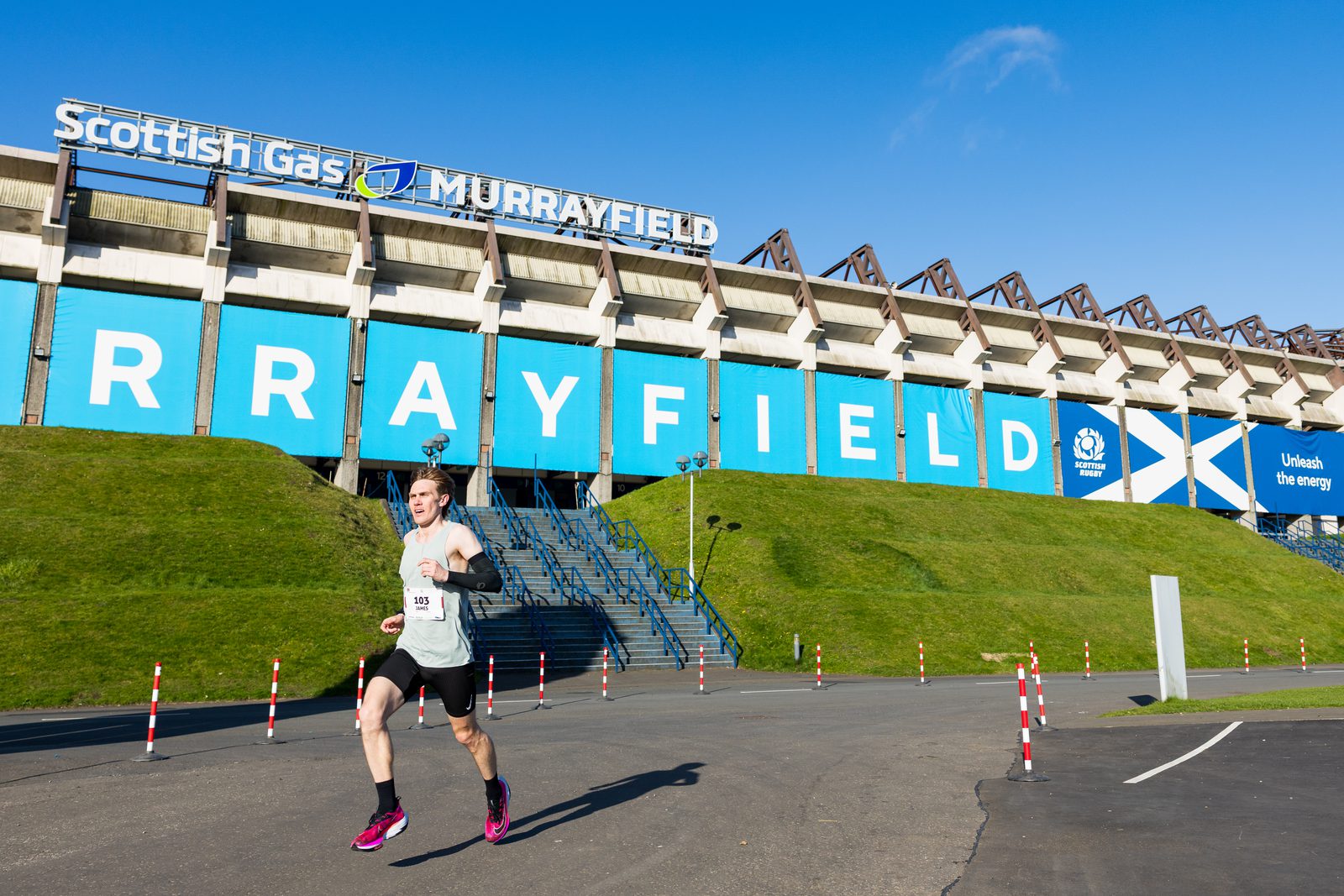 A runner wearing a race bib and athletic gear runs outside the Scottish Gas Murrayfield Stadium on a sunny day, with blue sky and stadium signage in the background.