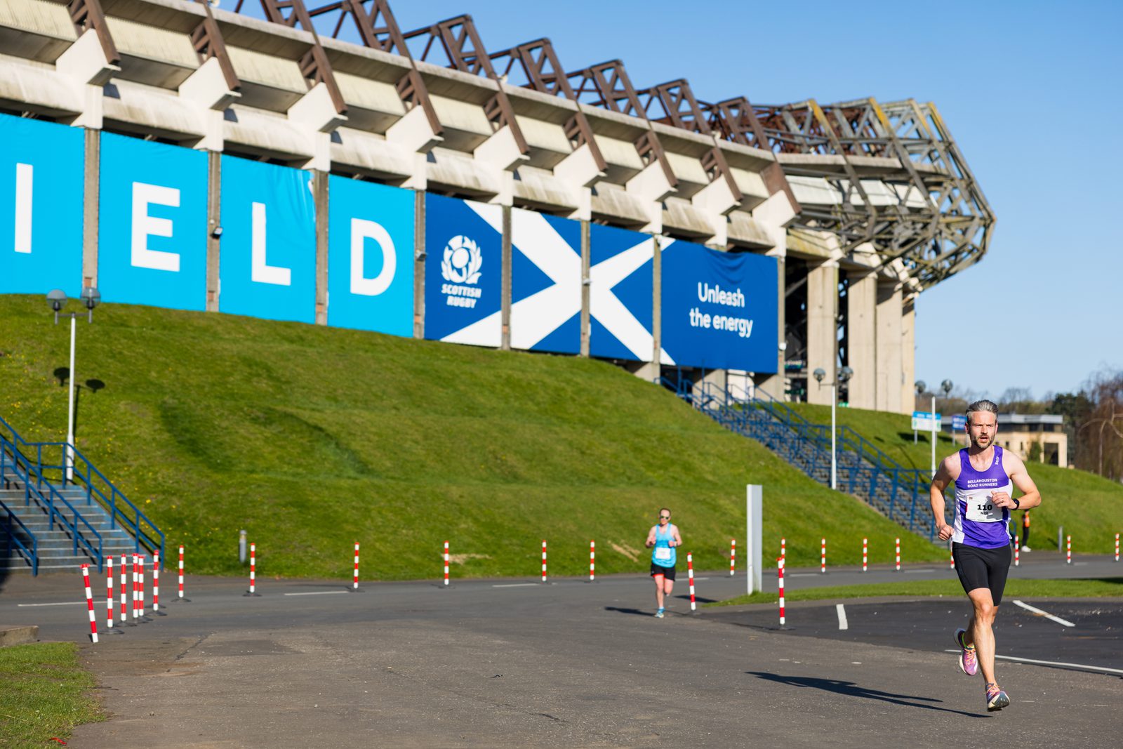 Two runners compete outside a large stadium with blue banners displaying the Scottish Rugby logo and the words "Unleash the energy." The sun is shining, and the sky is clear.