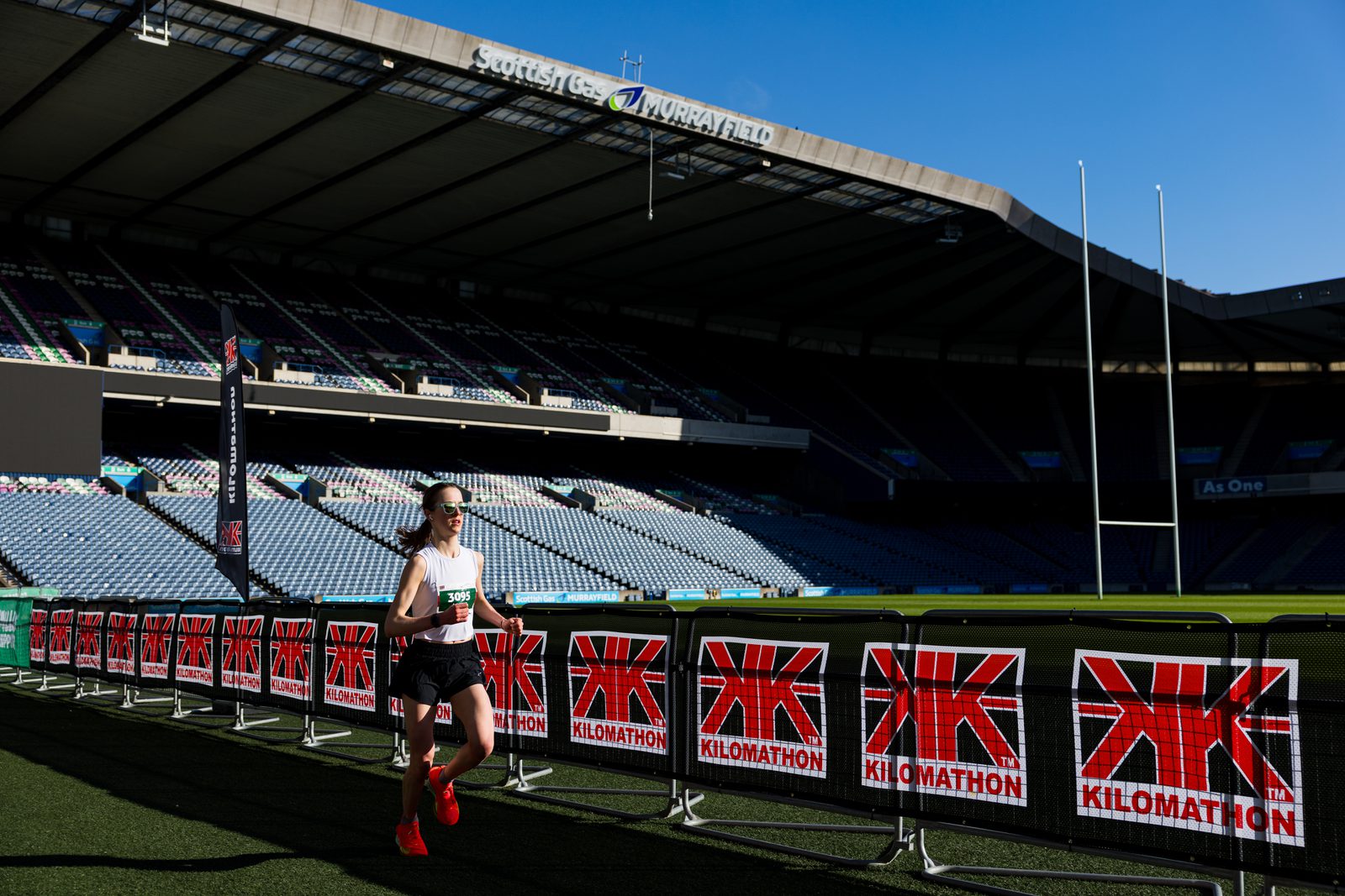 A runner in sunglasses and red shoes races past barriers with "KILOMATHON" signs inside a large, mostly empty stadium under clear blue skies.