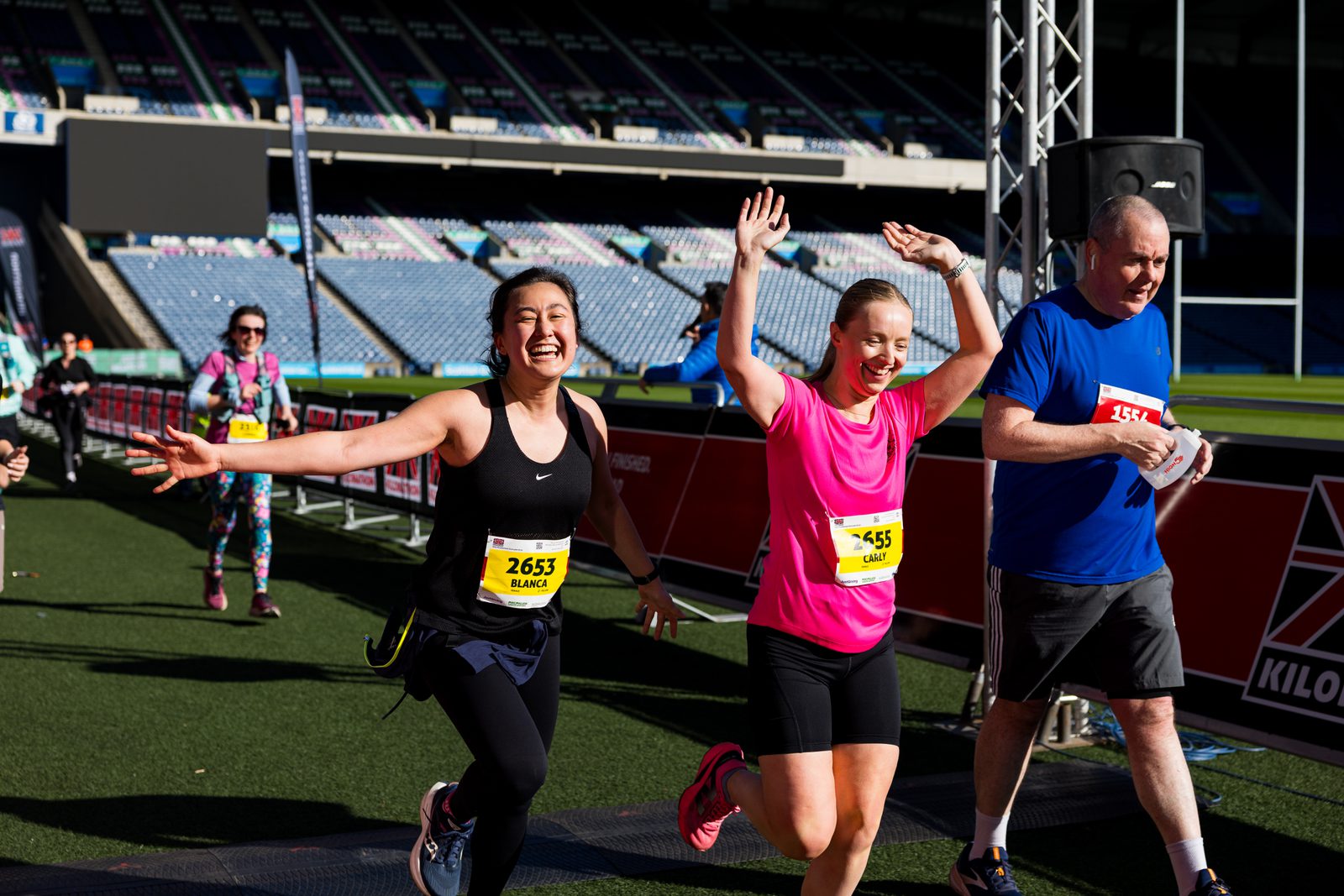 Two women with race bibs run joyfully toward the finish line, hands up and smiling, alongside a man. Other runners and empty stadium seats are visible in the background. The sun is shining brightly.