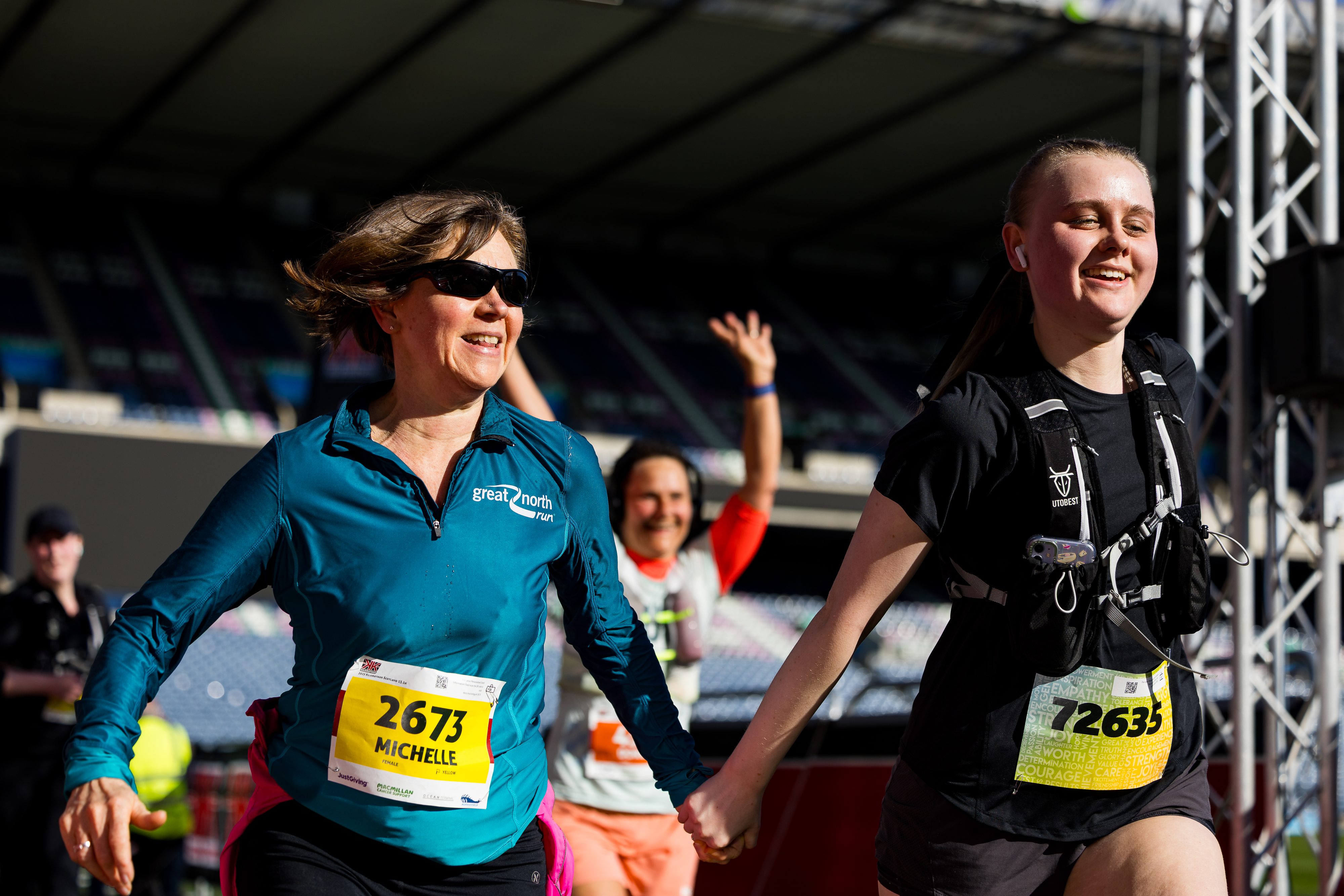 Two women with race bibs, smiling and holding hands as they run together in bright sunlight; a man behind them cheers with raised arms at an outdoor sporting event.