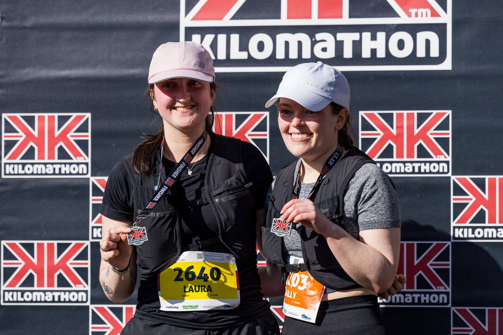 Two smiling runners wearing hats and holding medals pose in front of a KILOMATHON event backdrop. Both have race bibs visible, with numbers 2640 and 1403, and are dressed in athletic gear.