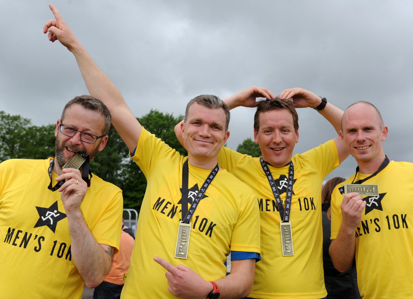 Four men in yellow "Men's 10K" shirts celebrate with medals. One bites his medal, another points upward, and all smile happily outdoors under a cloudy sky.