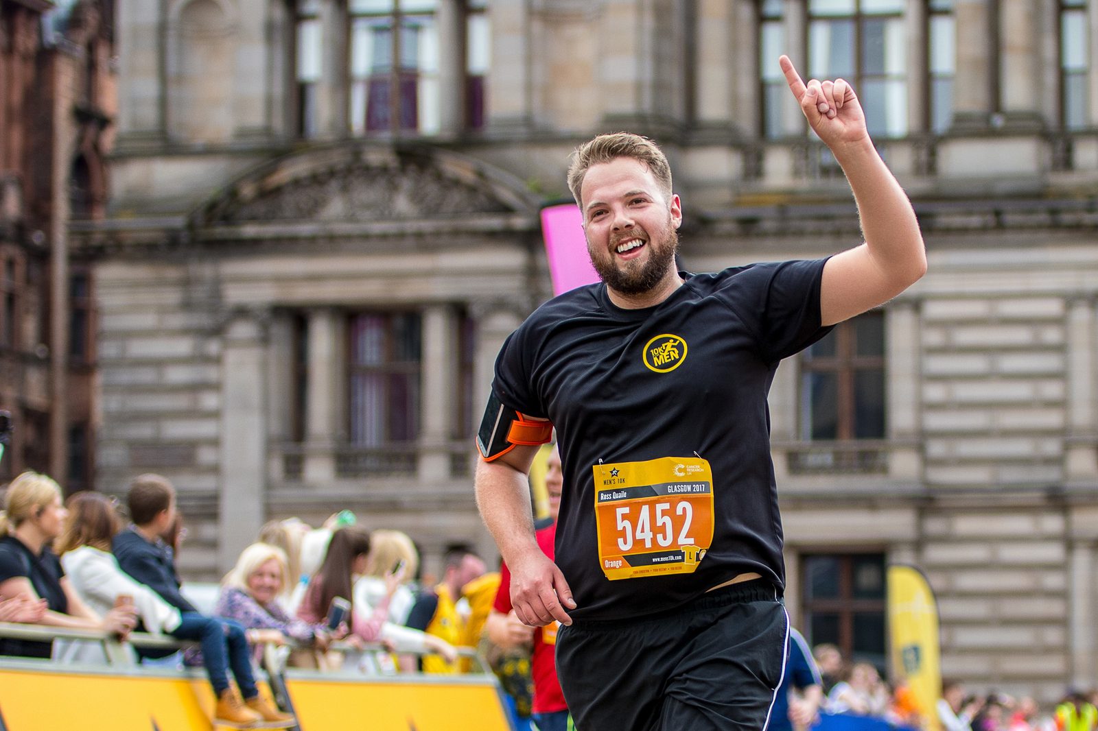 A man wearing a black shirt and race bib number 5452 raises his index finger in celebration as he runs past spectators during a race. Buildings with large windows are visible in the background, and the crowd is cheering him on.