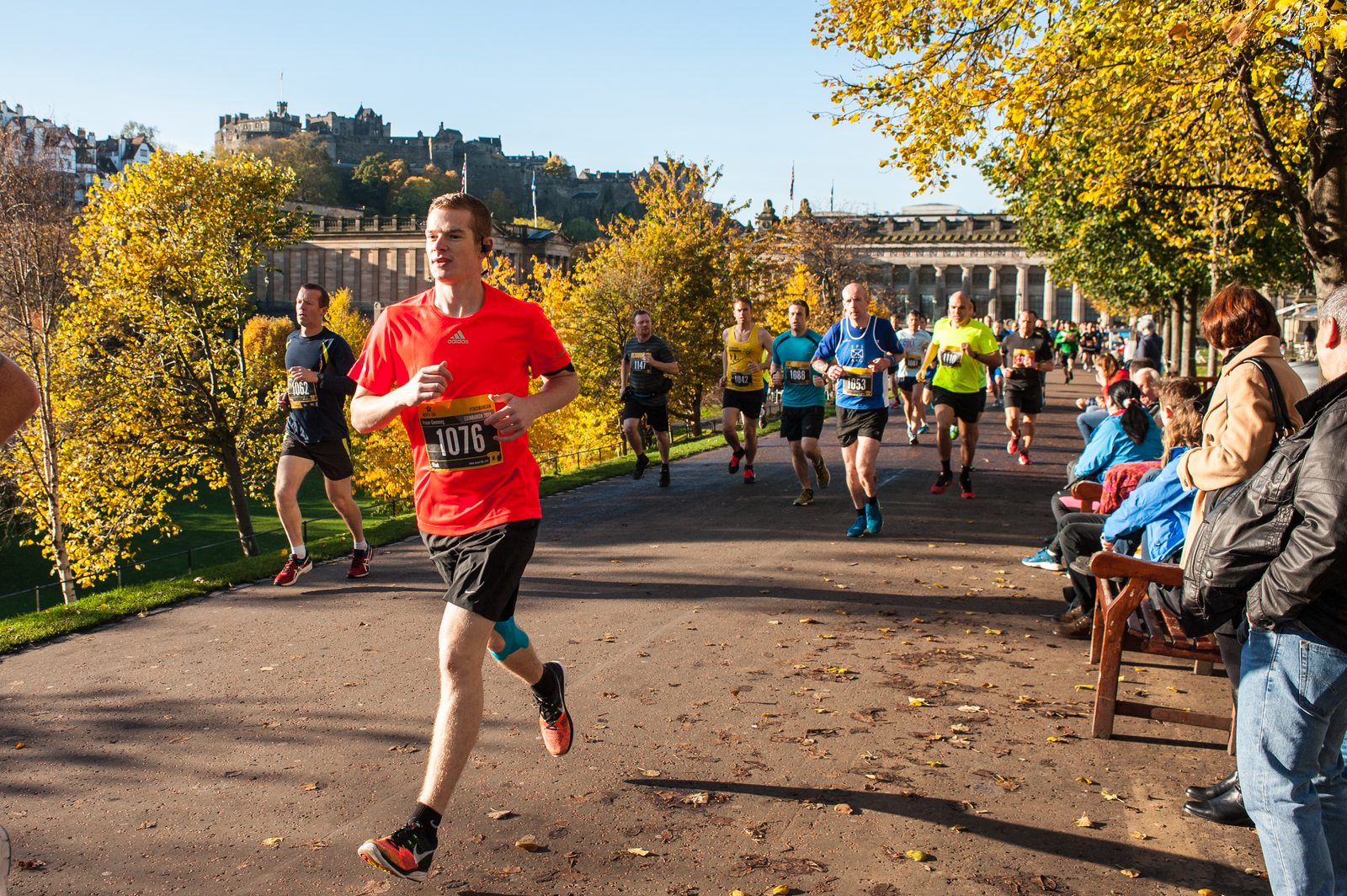 A group of runners participating in a race, with a man in a bright red shirt leading. Spectators sit and stand along the race path lined with autumn trees. A castle and historic buildings are visible in the background under a clear blue sky.