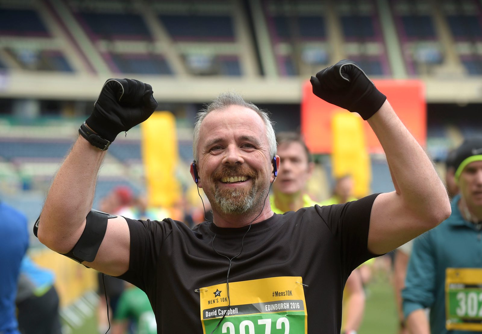A man wearing a black shirt and gloves raises his arms in triumph at the finish line of a race. He is laughing and has a race bib on his chest. Other runners and spectators can be seen in the background within a stadium.