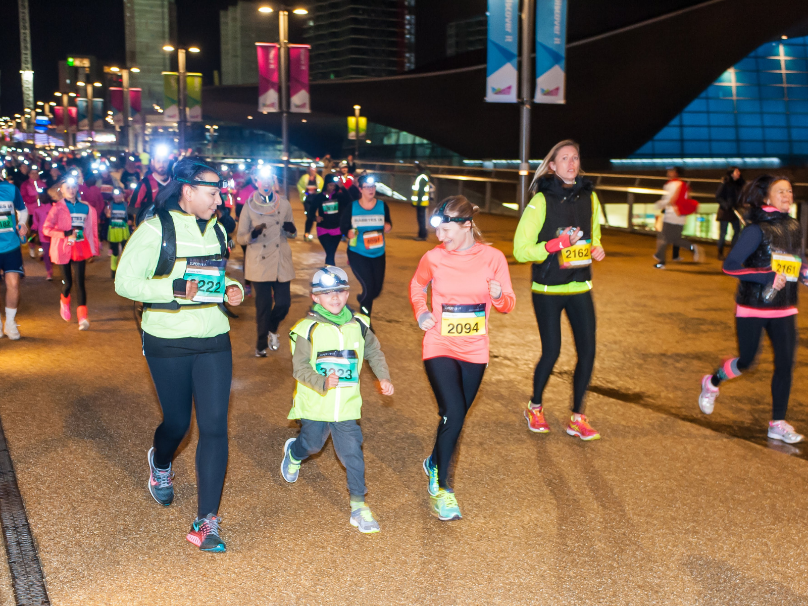 A group of runners, including adults and children, participate in a nighttime race. Most wear headlamps and bright athletic gear. Bib numbers are visible on their chests. The background shows lit buildings and signs indicating that the course is urban.
