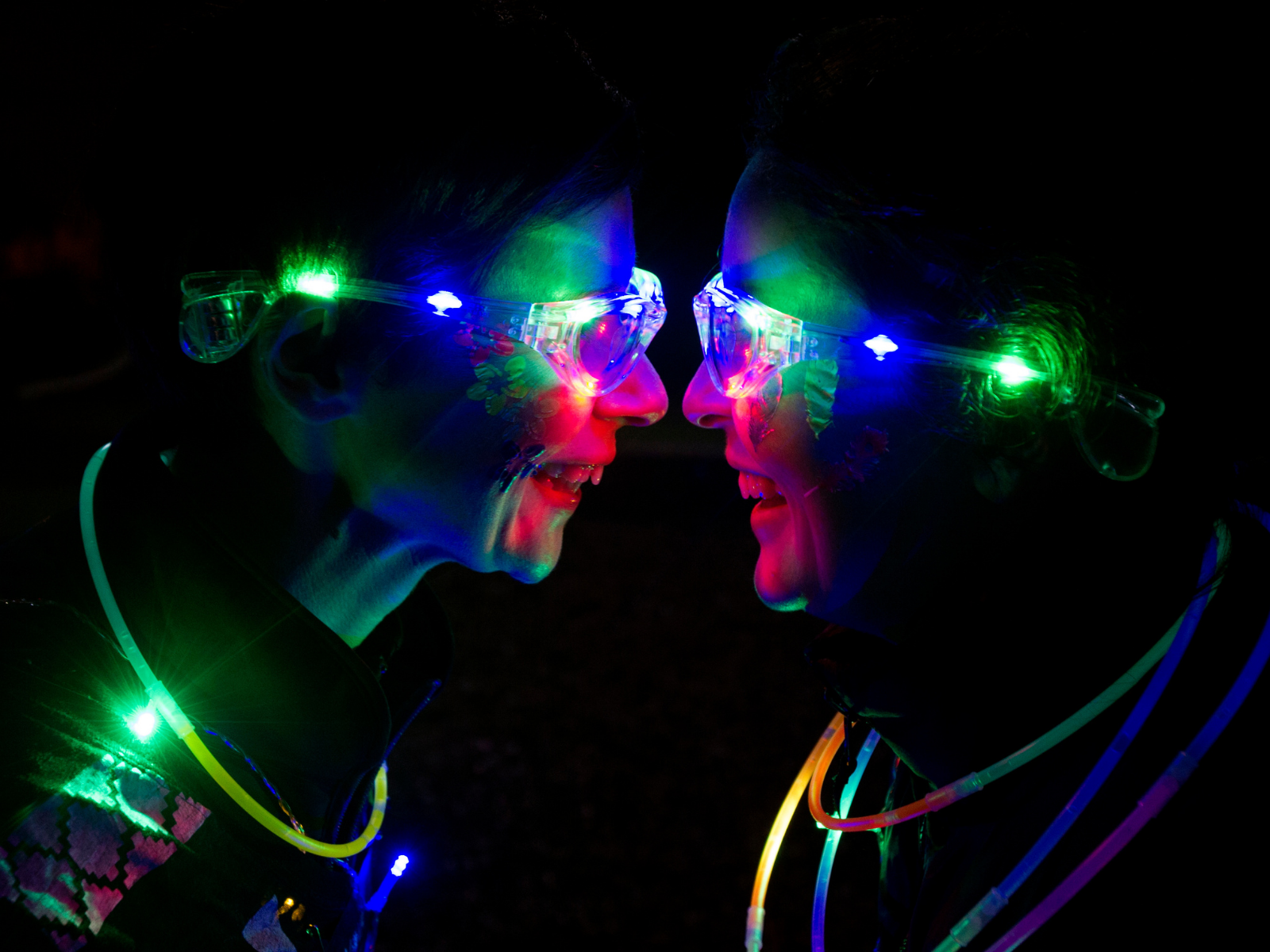 Two people face each other, smiling and laughing in a dark setting. They each wear glowing neon glasses and necklaces of different colors, including green, blue, and yellow, illuminating their faces with vibrant lights.