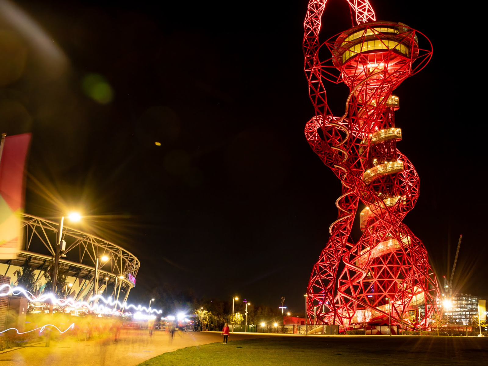 A nighttime view of the ArcelorMittal Orbit sculpture in London, brightly illuminated with red lights. To the left is part of the London Stadium. Blurred lights and people on the pathway suggest motion in the foreground. The sky is dark with a few stars visible.