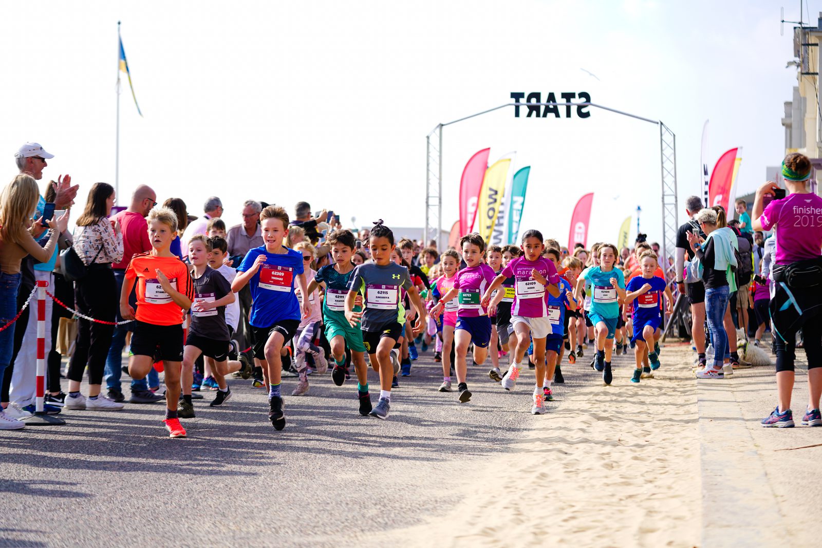 A group of children wearing colorful athletic gear are enthusiastically running at the start of a race. They are on a coastal pathway lined with spectators who are cheering them on. Various flags and race markers are visible in the background.